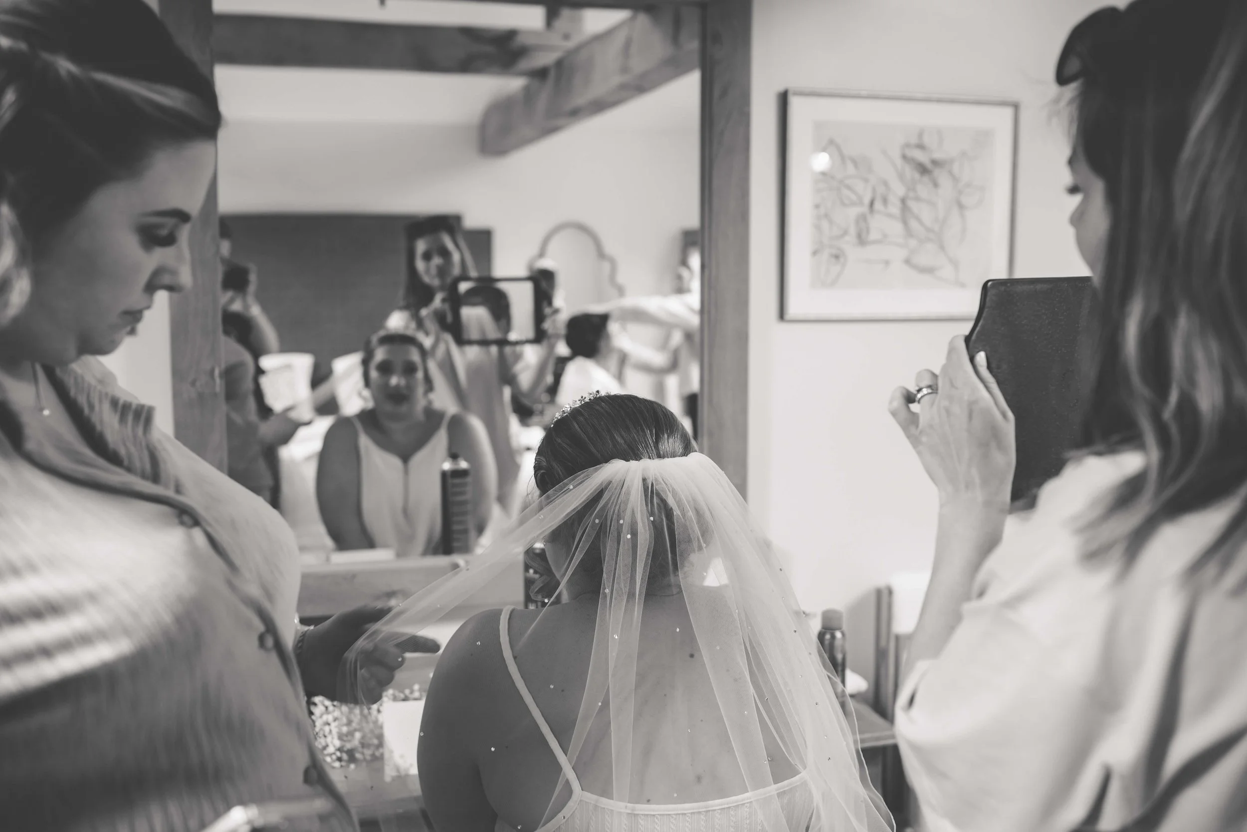 A bride is seated with her back to the camera, wearing a veil. Women around her assist and take photos, in a room with framed artwork on the walls, preparing for her wedding.