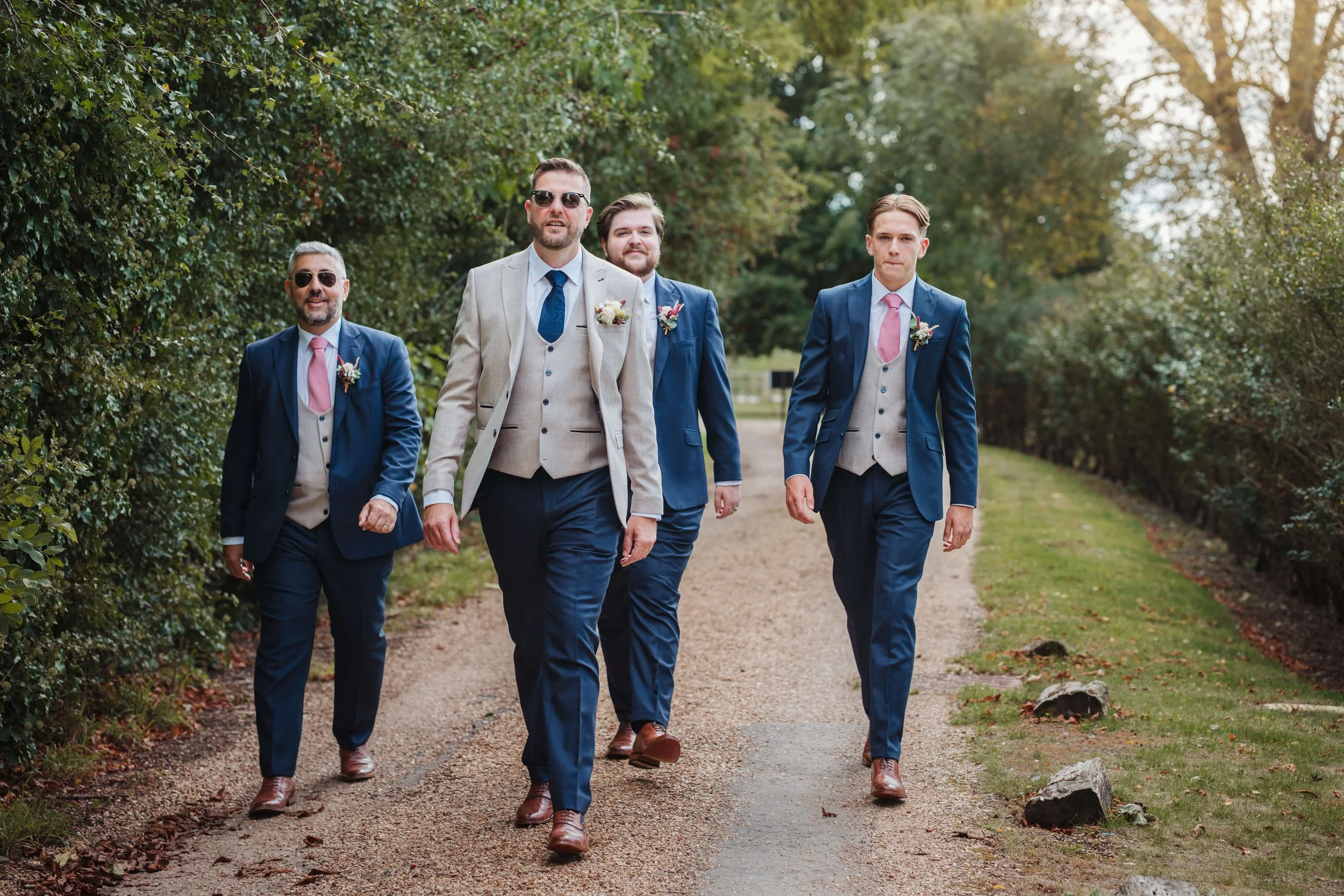 Five men dressed in suits walking on a gravel path in a green outdoor setting, likely at a wedding.