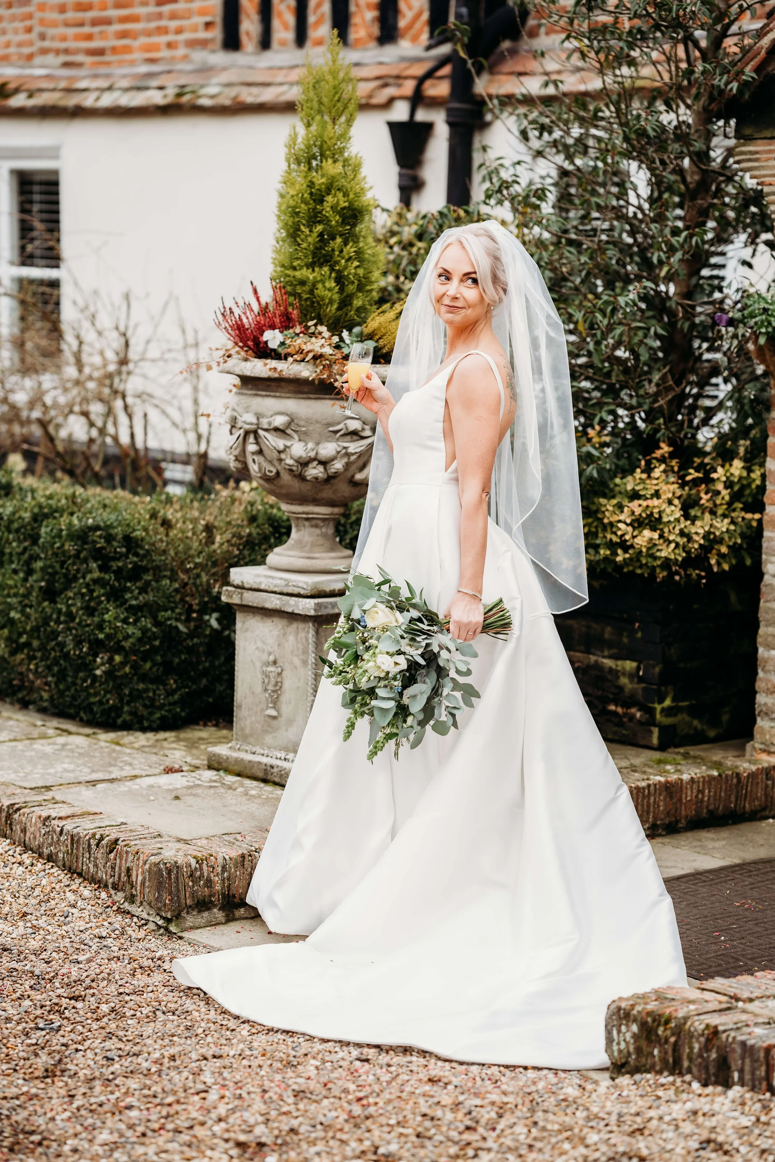 A bride in a white wedding dress holding a bouquet of white and green flowers, standing outdoors near a decorative stone planter with plants, and holding a glass of drink, smiling at the camera.