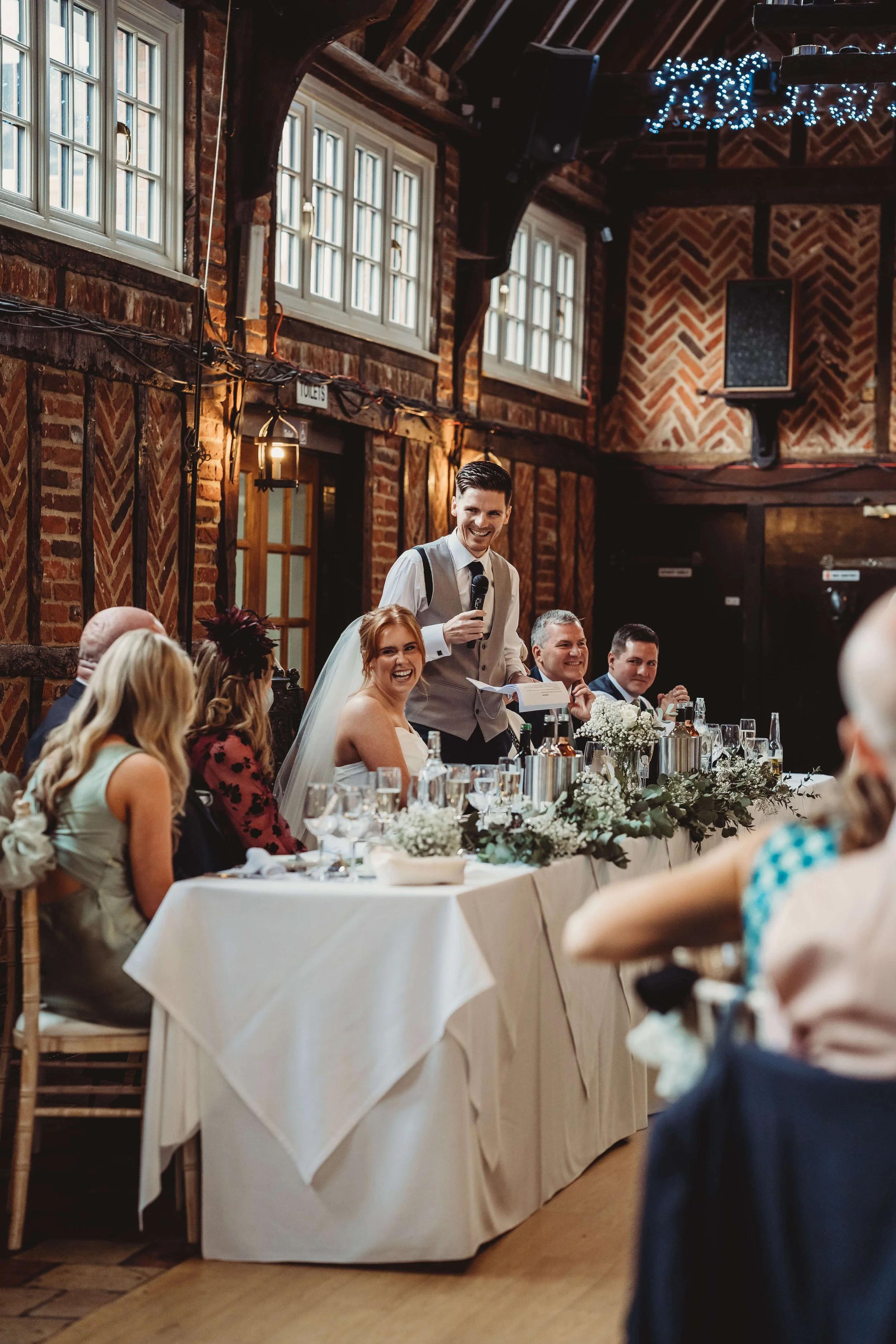 A wedding reception with a bride and groom sitting at a decorated table, giving a speech, with guests listening and smiling in a rustic venue with brick walls and large windows.