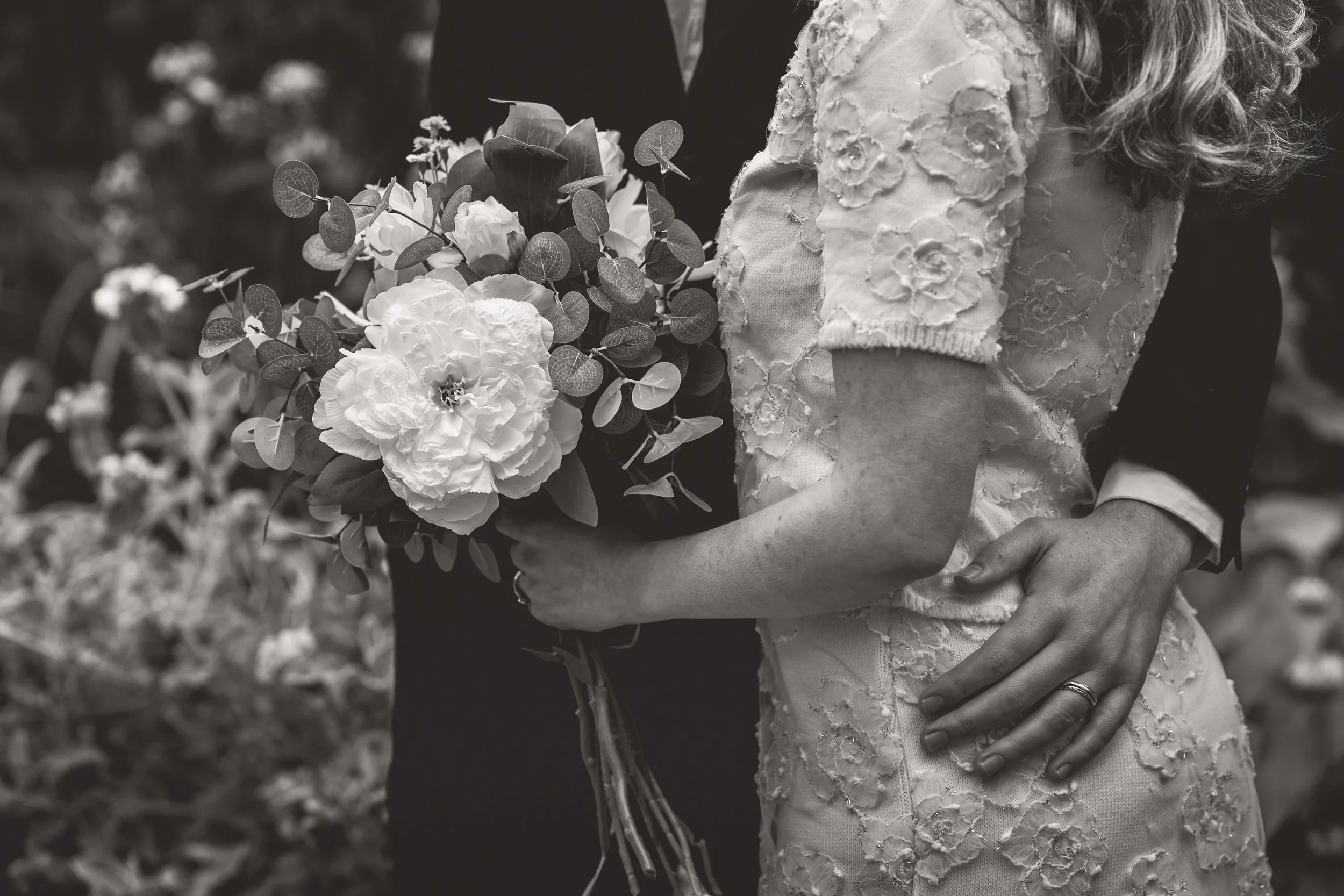 A bride holding a bouquet of flowers, standing close to a groom, with her hand resting on her waist. The image is in black and white.