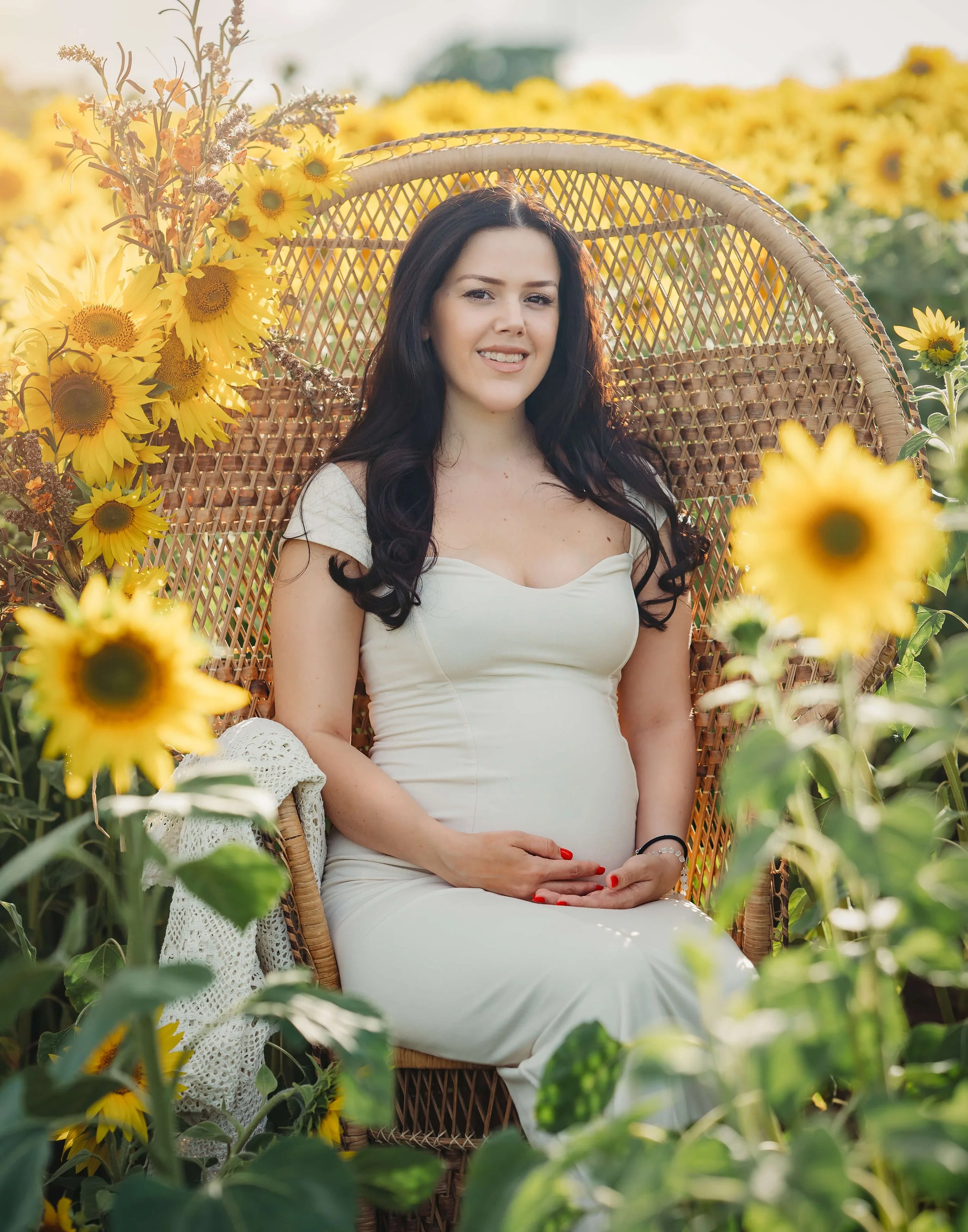 A woman in a cream dress sitting on a wicker chair surrounded by sunflowers in a sunflower field during golden hour.