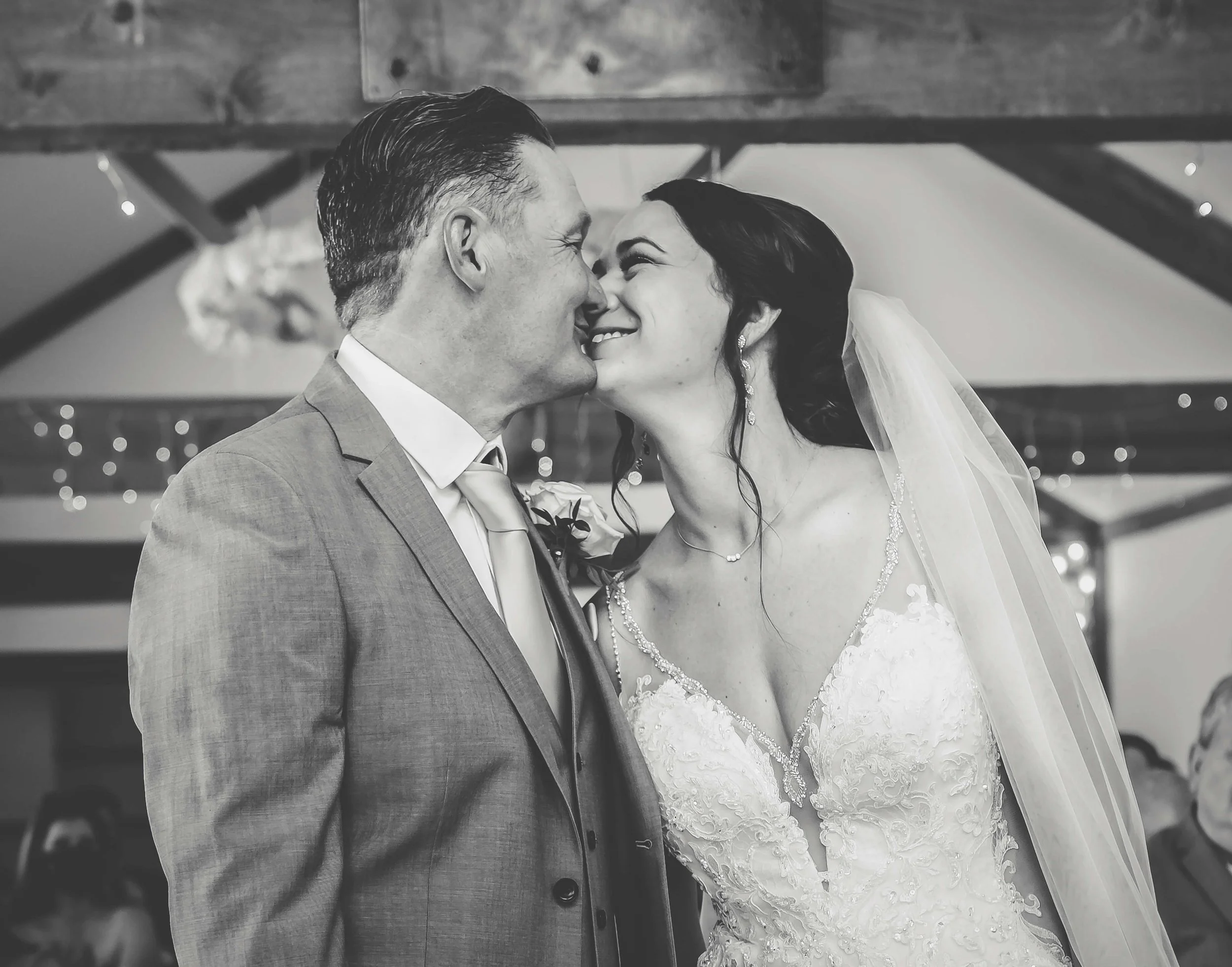 Black and white photo of a bride and groom touching noses during wedding, smiling with close eyes, indoor decorated setting.