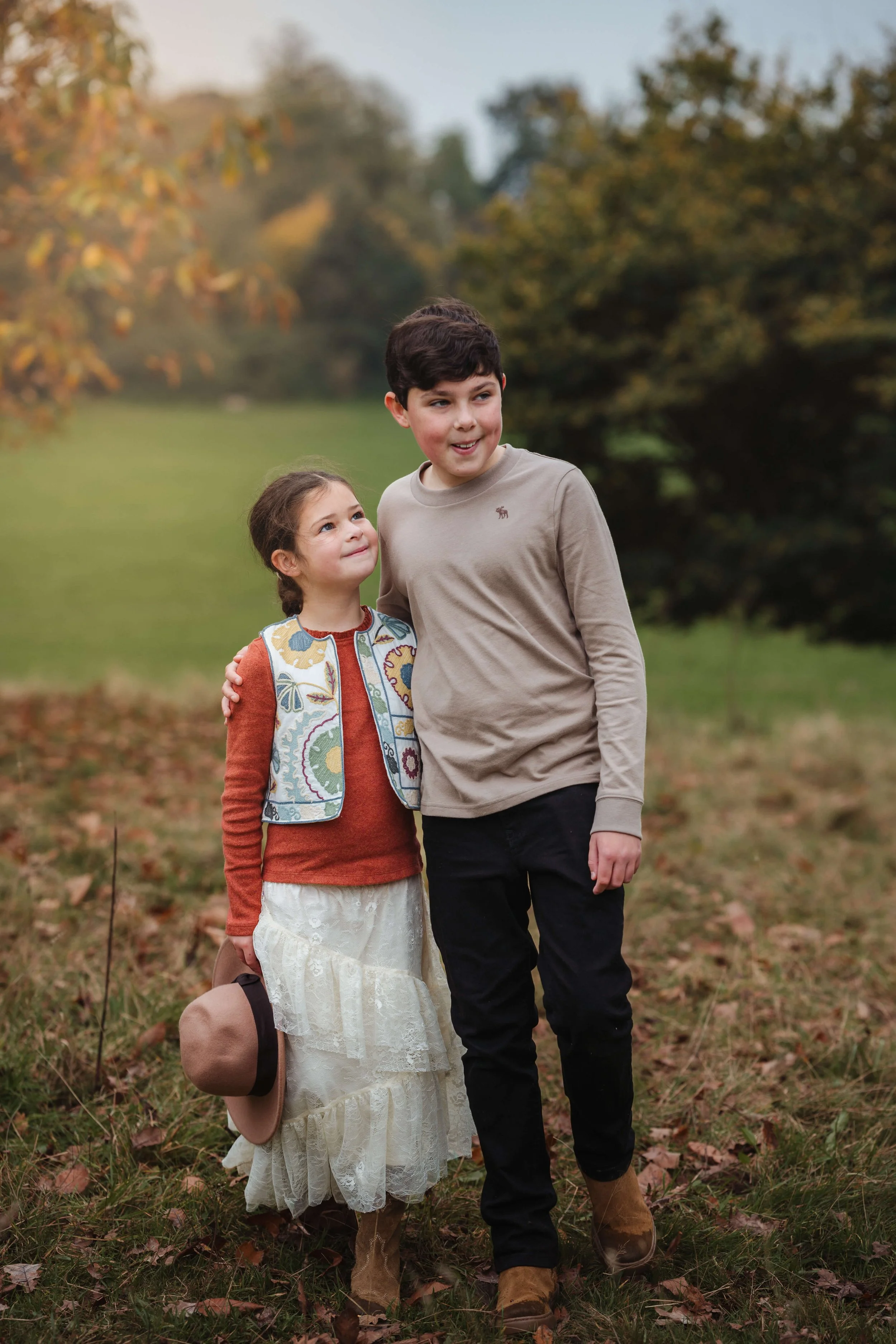 A young girl and a teenage boy walking together outdoors in a park during autumn. The girl is holding a hat and wearing a colorful vest, while the boy has his arm around her shoulder and is dressed in a light-colored long sleeve shirt and dark pants.