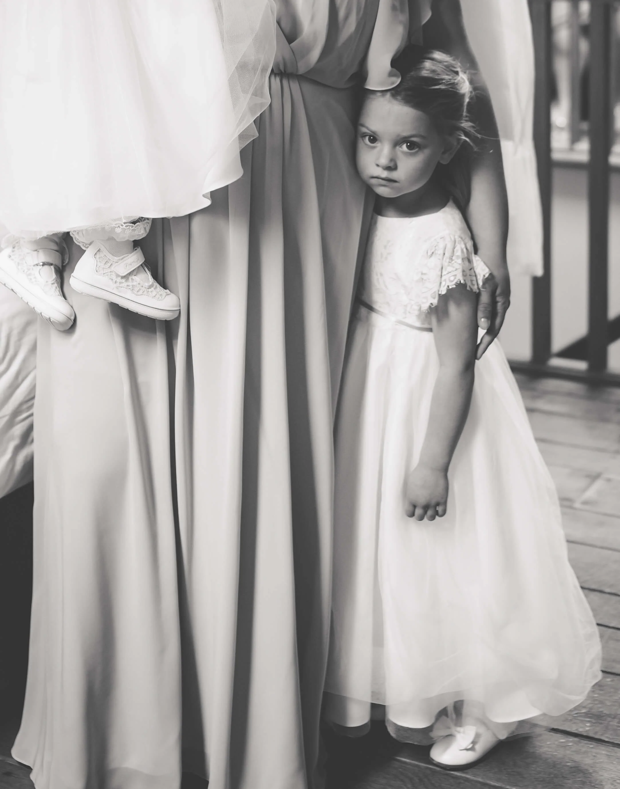 A young girl in a white dress holding her arm around an adult's waist, with a serious expression, standing next to the adult who is dressed in a gown, part of which is visible, and holding a child in white shoes.