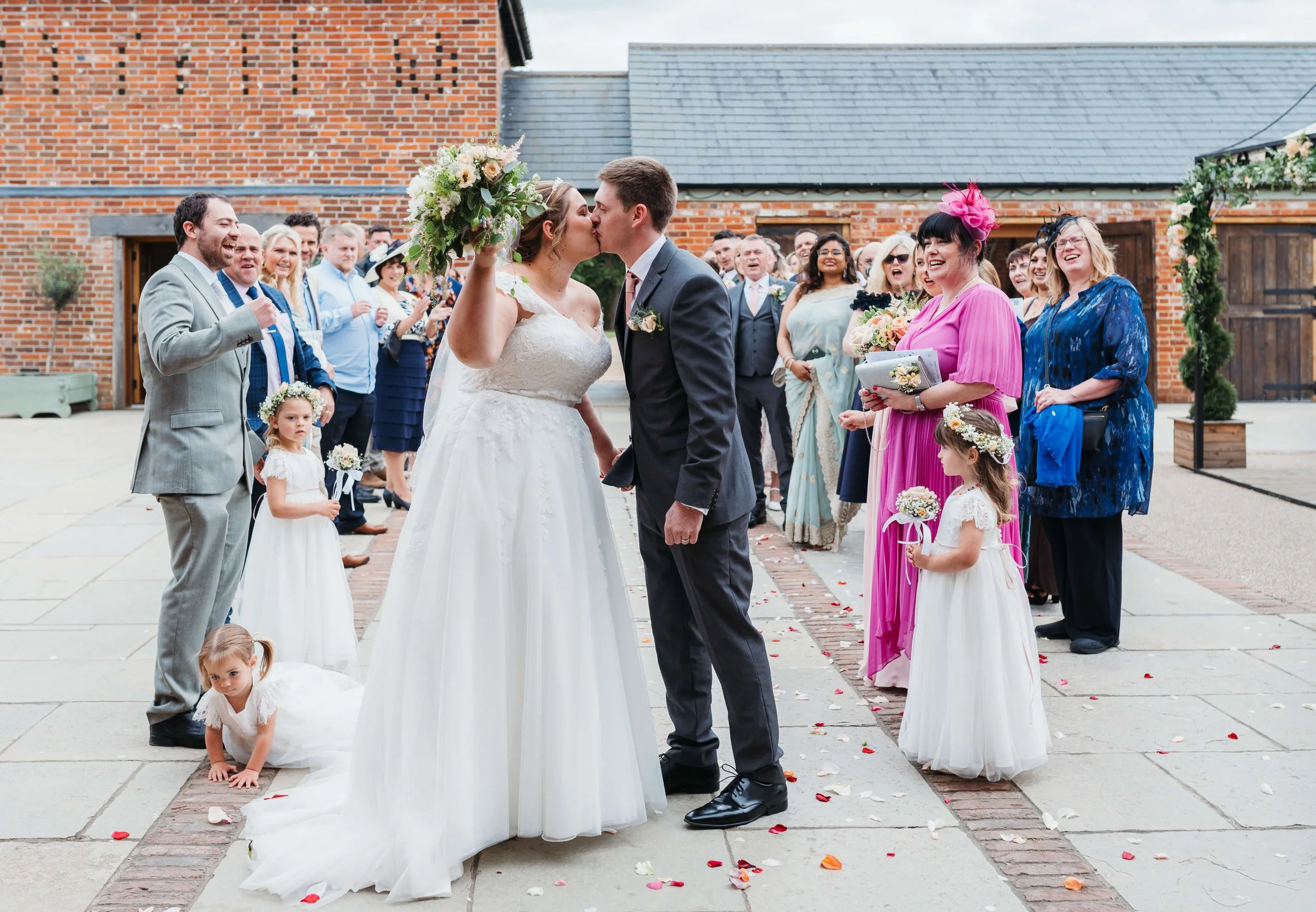 Bride and groom sharing a kiss at their wedding ceremony outdoors, surrounded by guests, bridesmaids, and flower girls.