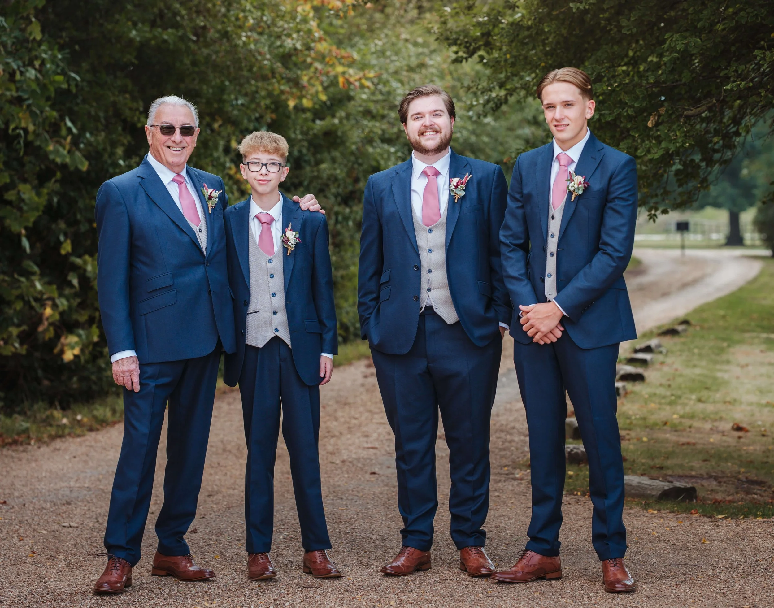 Five men dressed in matching blue suits with pink ties, standing outdoors on a gravel path surrounded by greenery, likely at a wedding or formal event.