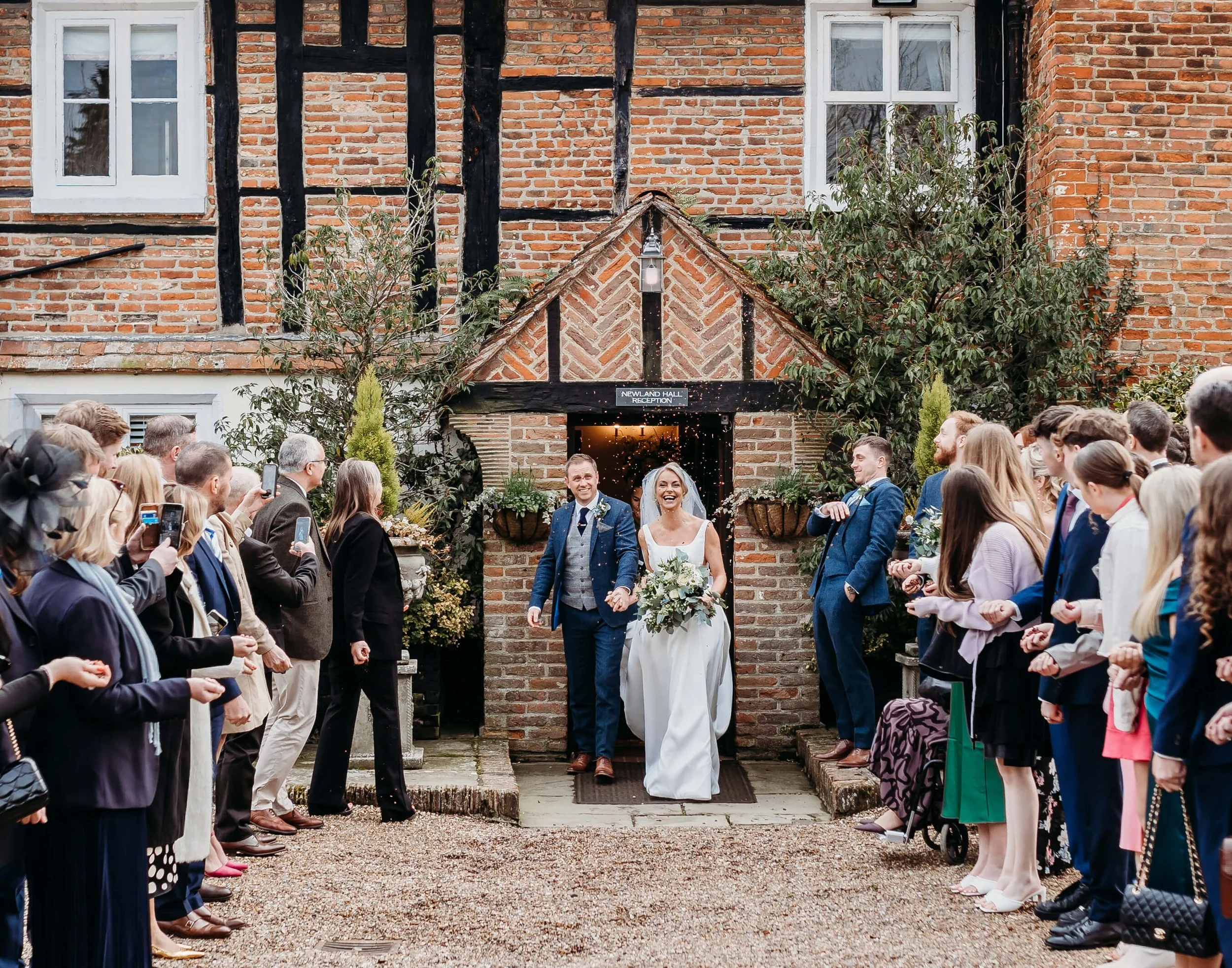 Happy bride and groom walking out of a brick building, surrounded by wedding guests taking photos and cheering.