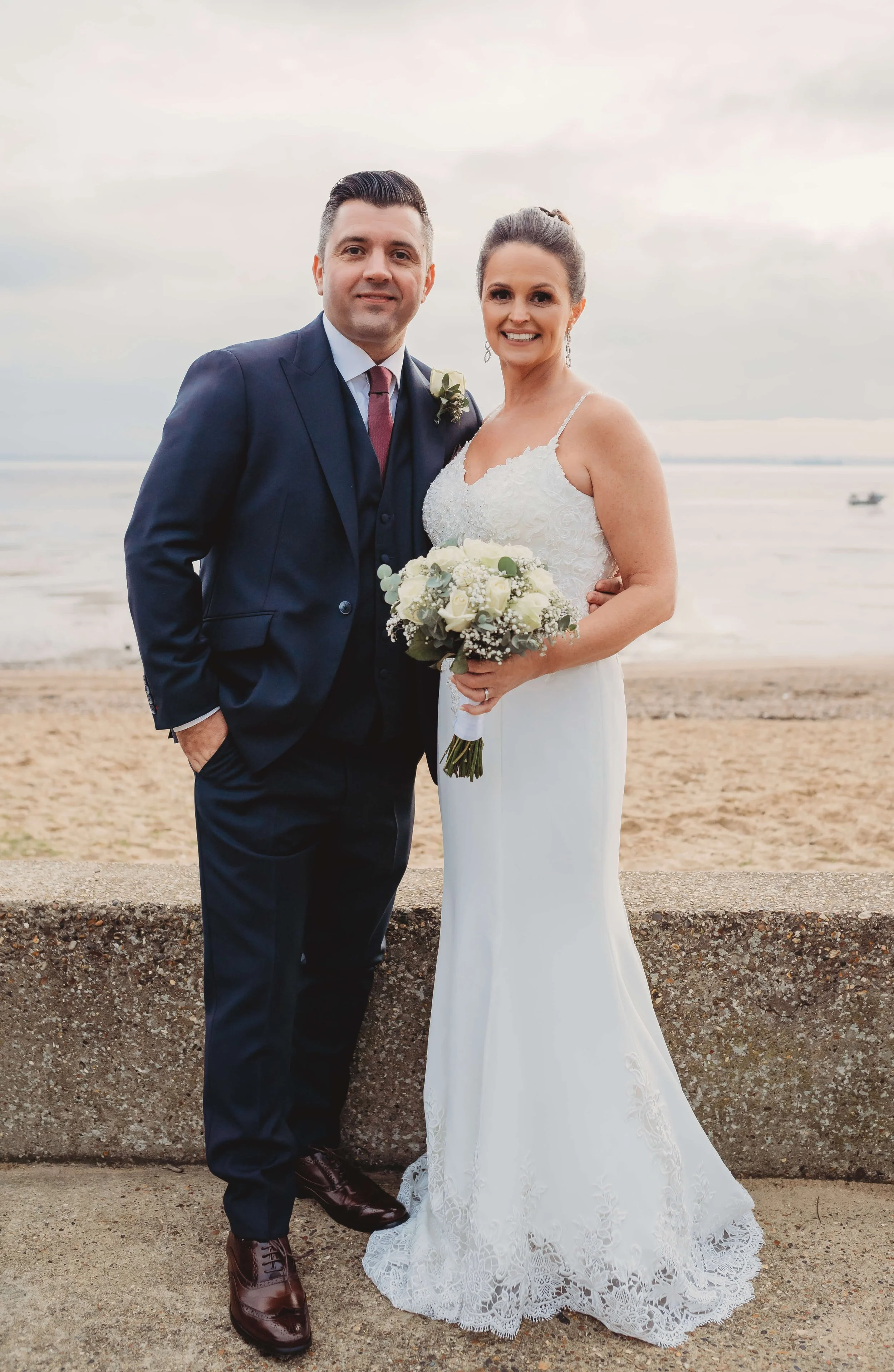 A bride and groom standing together on a beach, dressed in wedding attire. The bride is holding a bouquet of white roses and greenery, and the groom has a boutonniere on his suit lapel. The couple is smiling at the camera, with the ocean and a cloudy