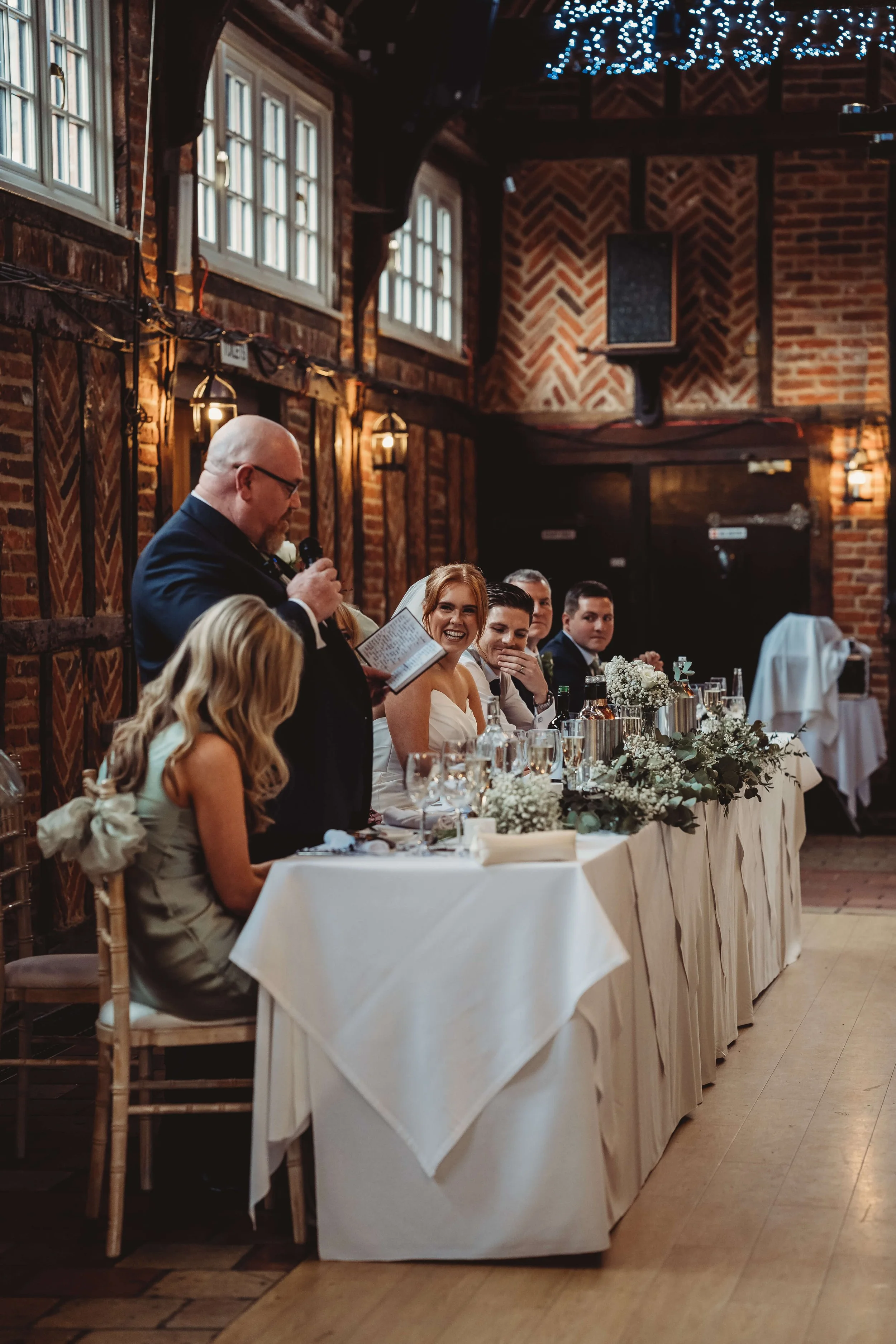 A wedding reception with a long table, floral decorations, and a man giving a toast while the guests listen and smile.