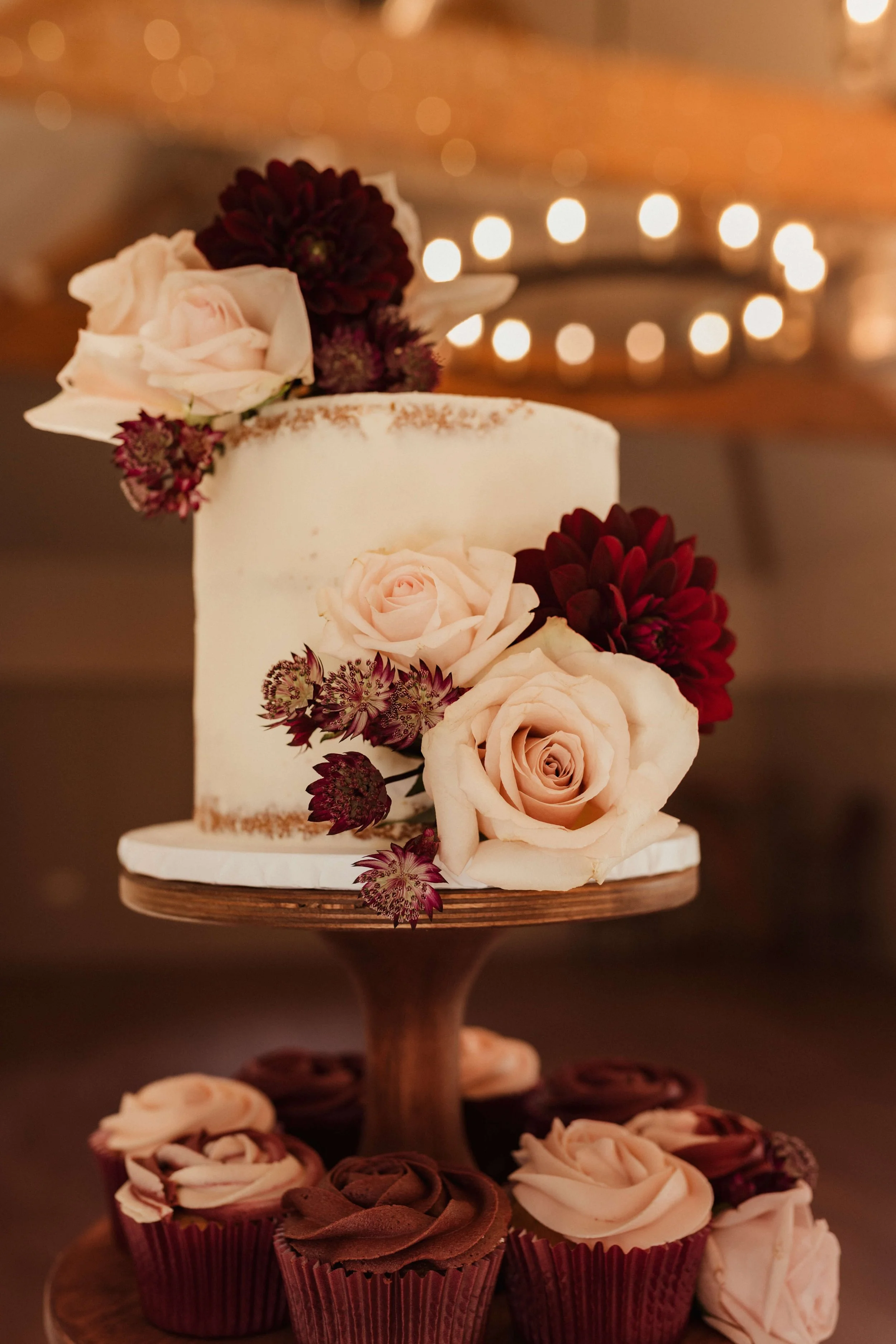 A wedding cake decorated with light pink roses and dark red flowers, displayed on a wooden cake stand surrounded by chocolate and pink frosting cupcakes.