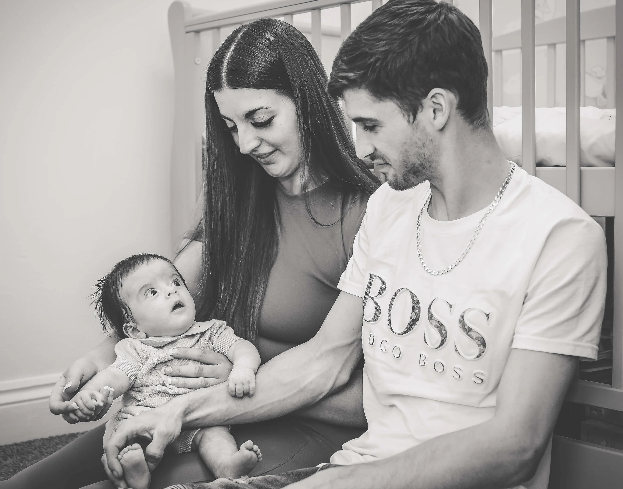 A black and white photo of a family in a nursery. The mother is holding a surprised-looking baby girl, while the father sits nearby, smiling at the child.