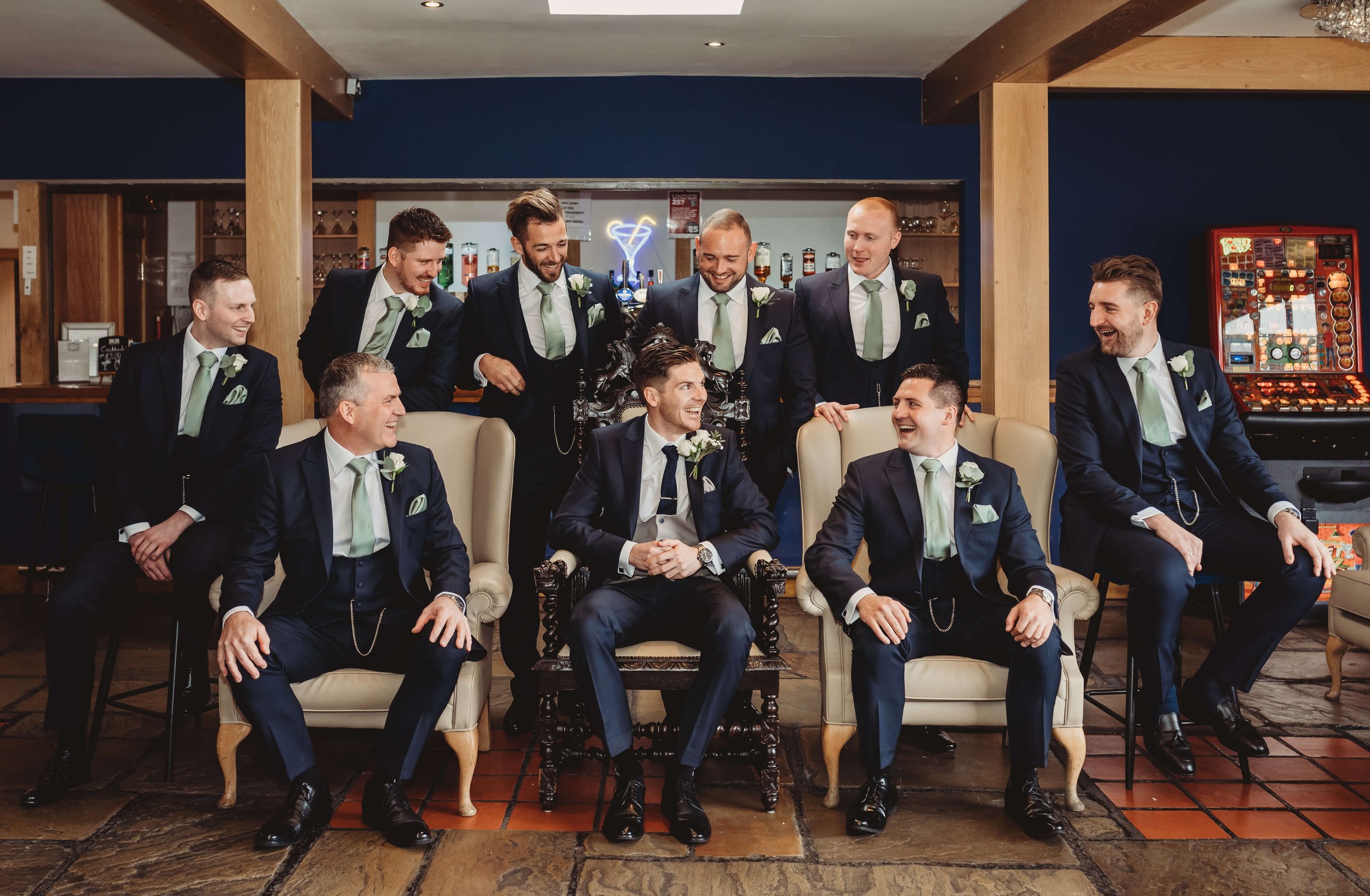 A group of groomsmen in suits, some seated in chairs and others standing behind, smiling and looking at each other in a social setting, possibly a bar or lounge.