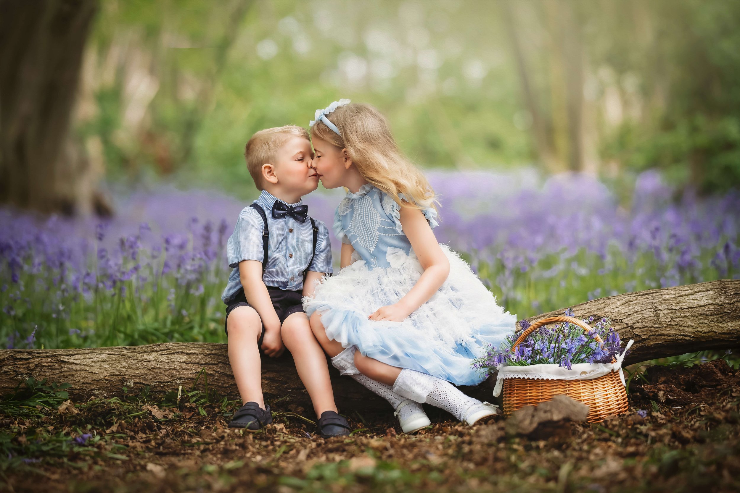 Two young children, a boy and a girl, sitting on a log in a forest, leaning in to kiss each other. The girl is dressed in a blue and white dress, the boy in a blue shirt with a bow tie. There is a basket of purple flowers beside them.