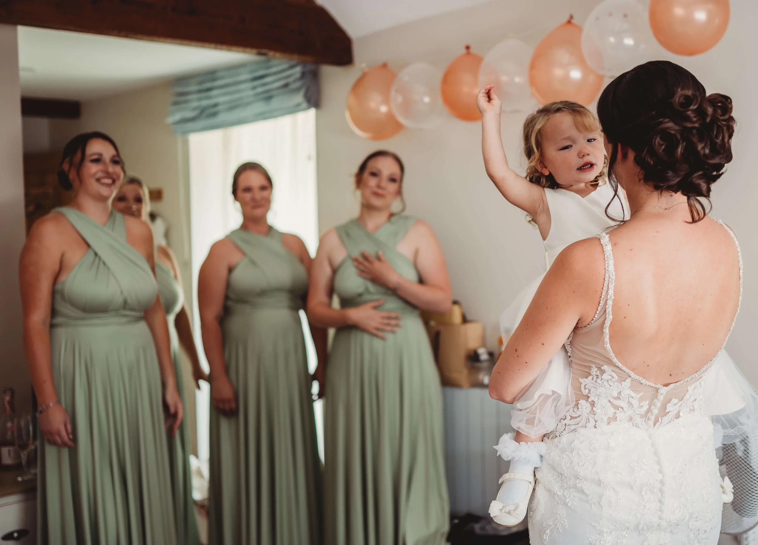 A bride holding a young girl with curly blond hair, wearing a white dress, is surrounded by four women in matching light green dresses, with a group of peach and clear balloons hanging from the ceiling.