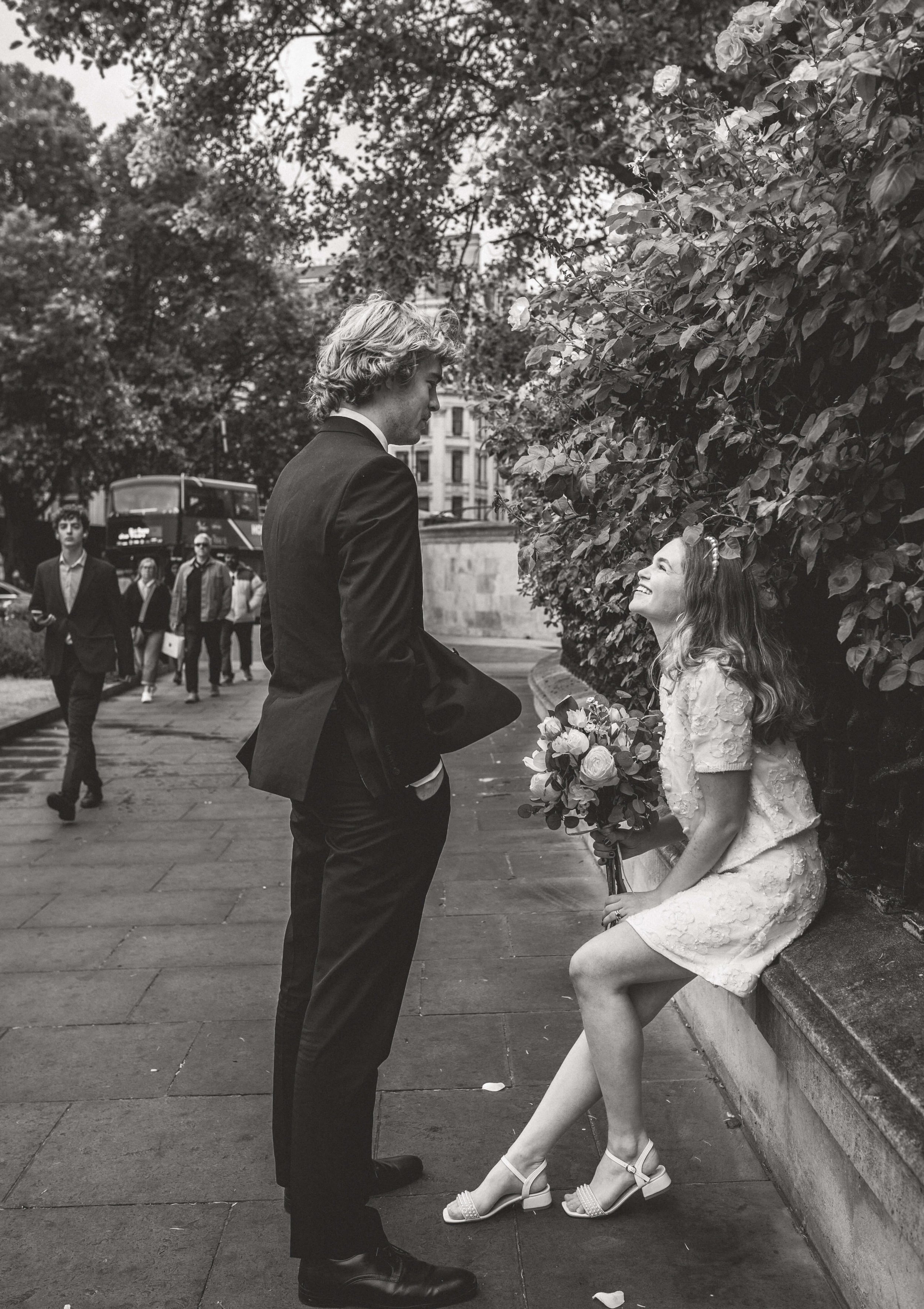 A black-and-white photo of a young woman sitting on a stone bench with a dress and heels, holding a bouquet of flowers, and smiling at a tall young man in a suit standing in front of her on a city sidewalk.