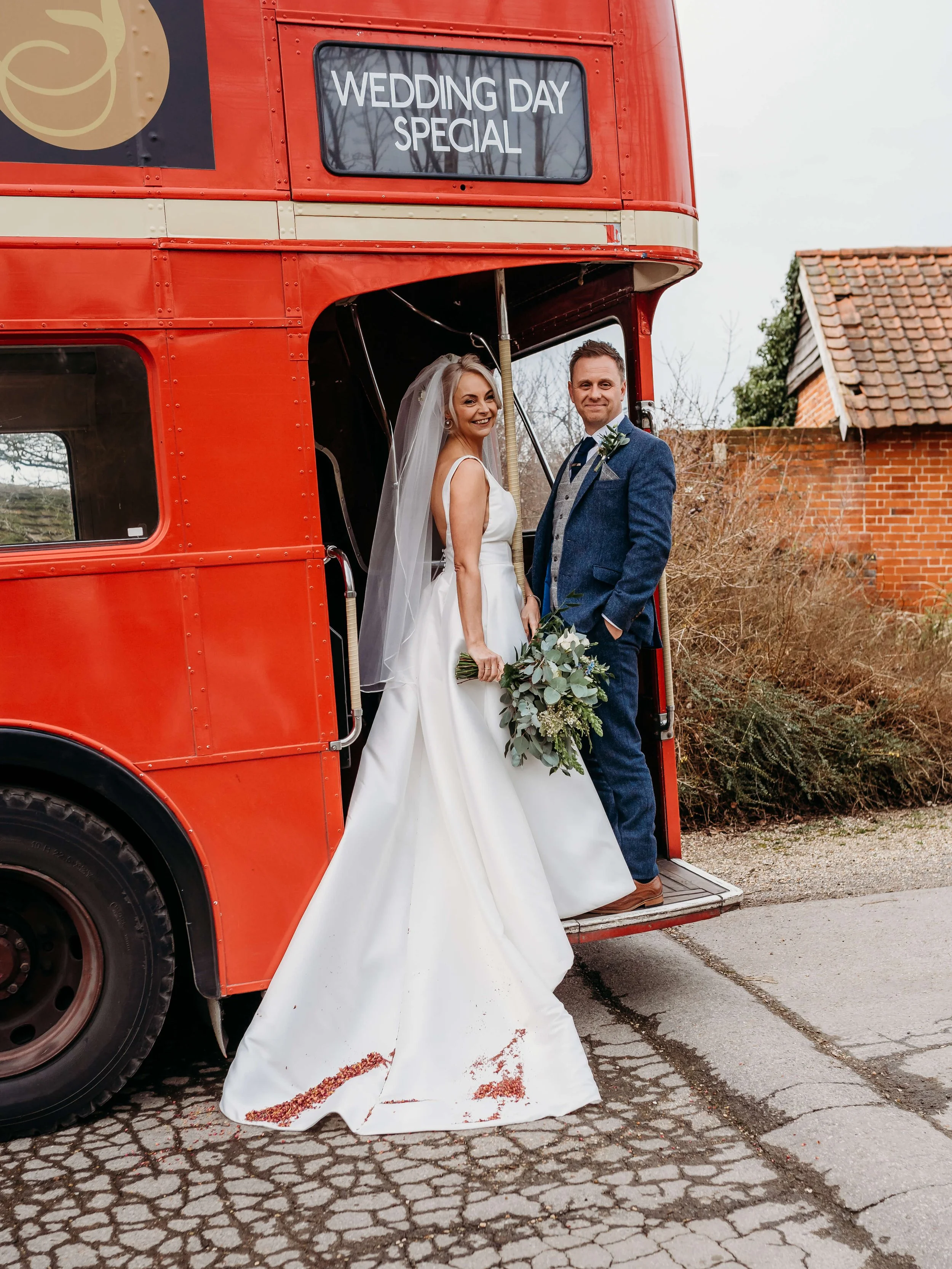 Bride and groom standing on a classic red double-decker bus, with a sign reading 'Wedding Day Special' on top, outside near a house and shrubs.