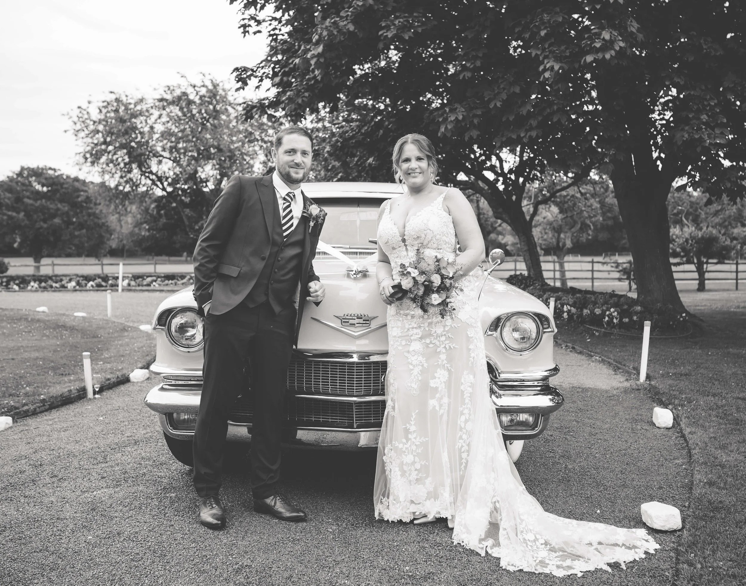 Black and white photo of a bride and groom standing in front of a vintage car on a wedding day. The groom is wearing a dark suit with a striped tie, and the bride is in a lace wedding gown holding a bouquet. Trees and a fence are in the background.