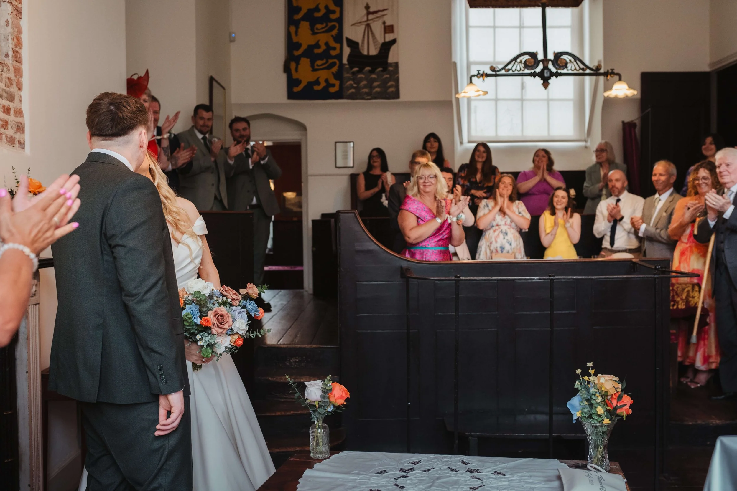 A bride and groom stand in front of a gathering of wedding guests in a decorated indoor venue, with guests applauding and smiling.