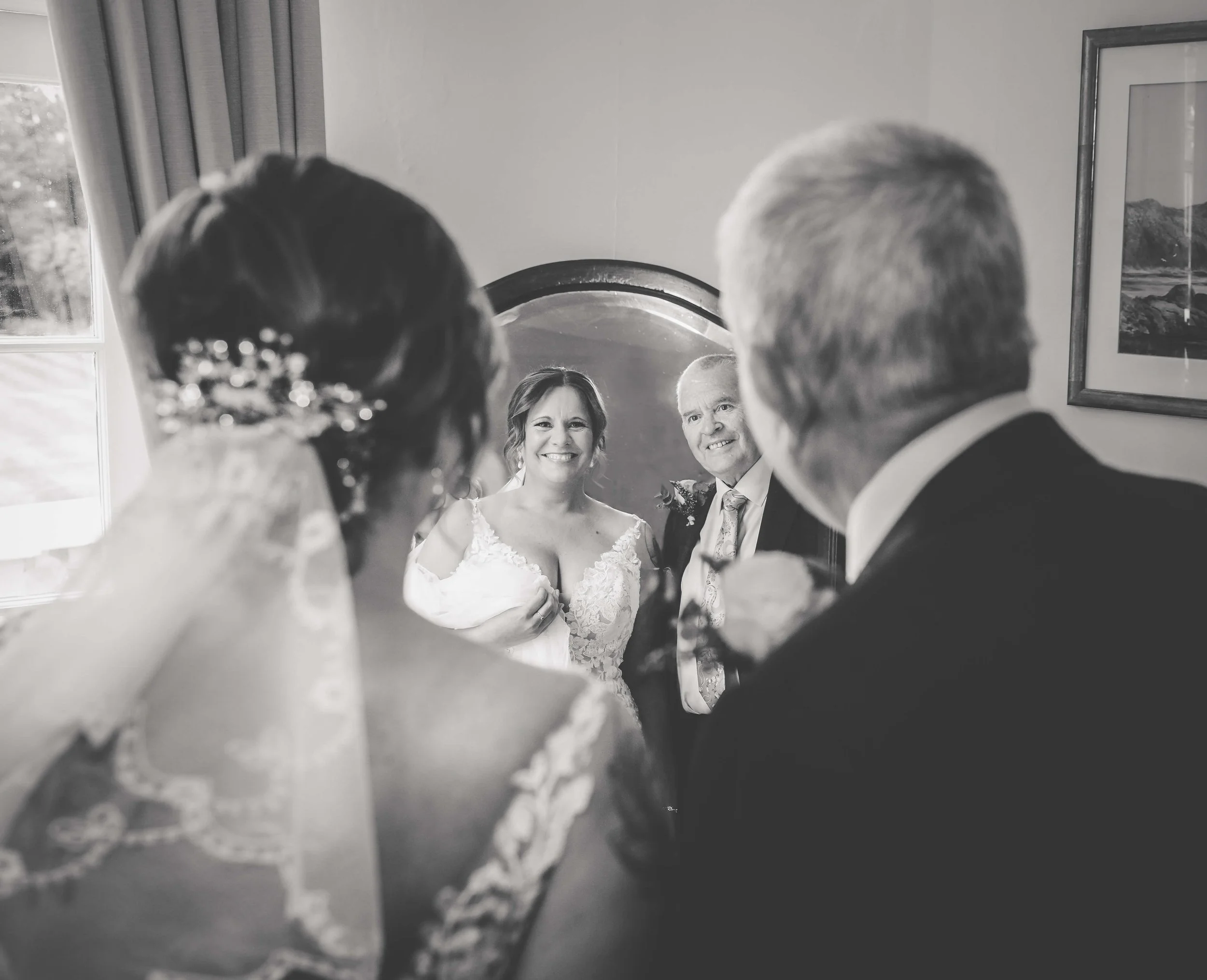 A bride and groom standing in front of a mirror, smiling at each other, with the reflection of a guest, possibly the bride's mother, smiling back. The scene is in a room with a window and framed pictures on the wall.