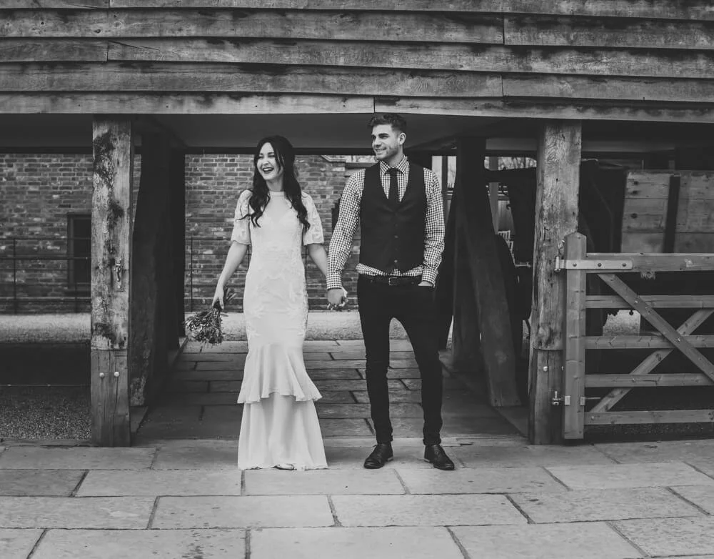 A couple holding hands and walking under a rustic wooden structure outdoors, smiling and dressed in wedding attire. The woman wears a long white wedding dress, and the man wears a checkered shirt with a vest and dark pants.