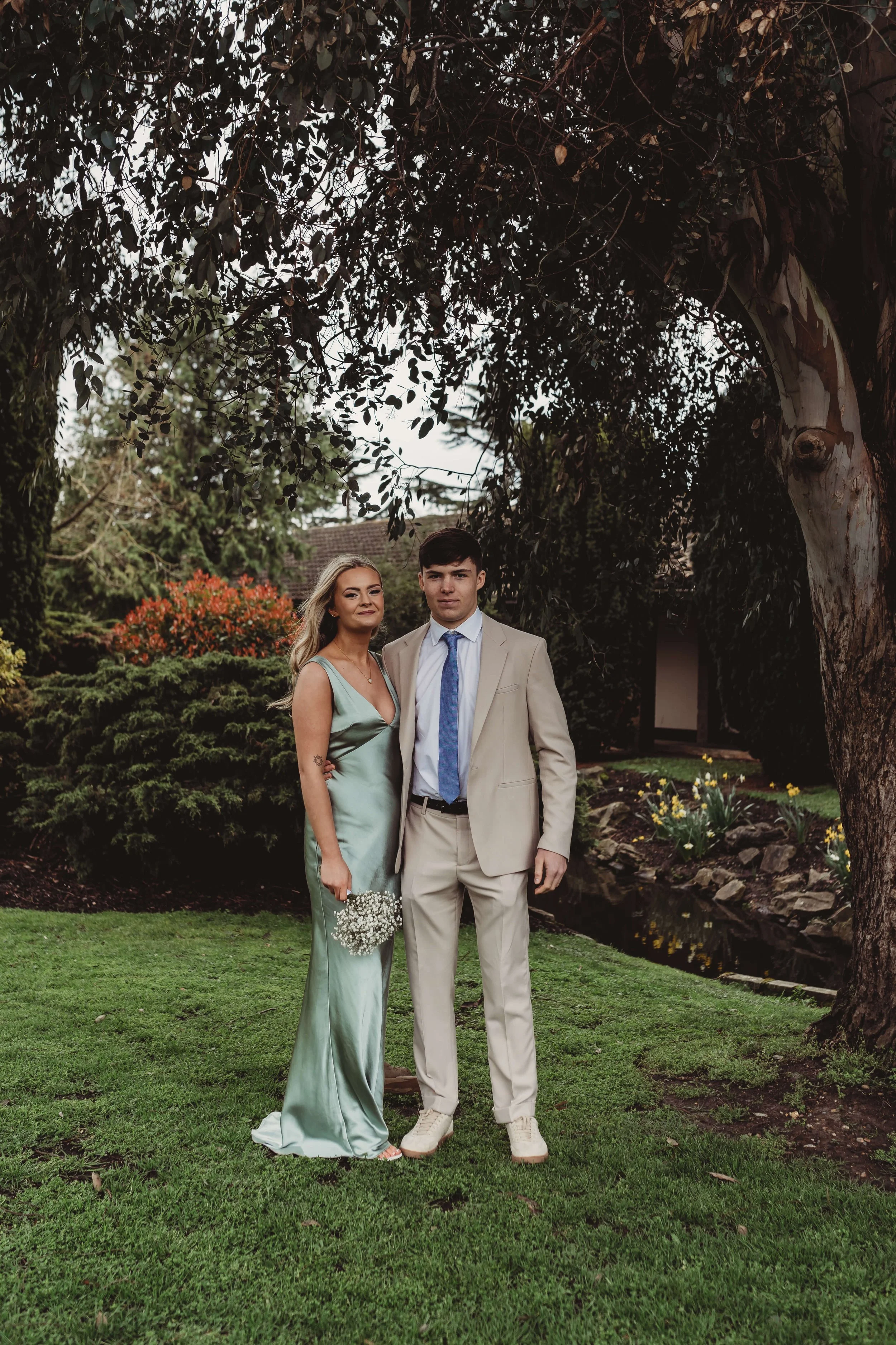 A young man and woman stand together outdoors under a tree. The woman wears a long, light green satin dress and holds a small bouquet of white flowers. The man wears a beige suit with a white shirt and a blue tie. The background features lush green b