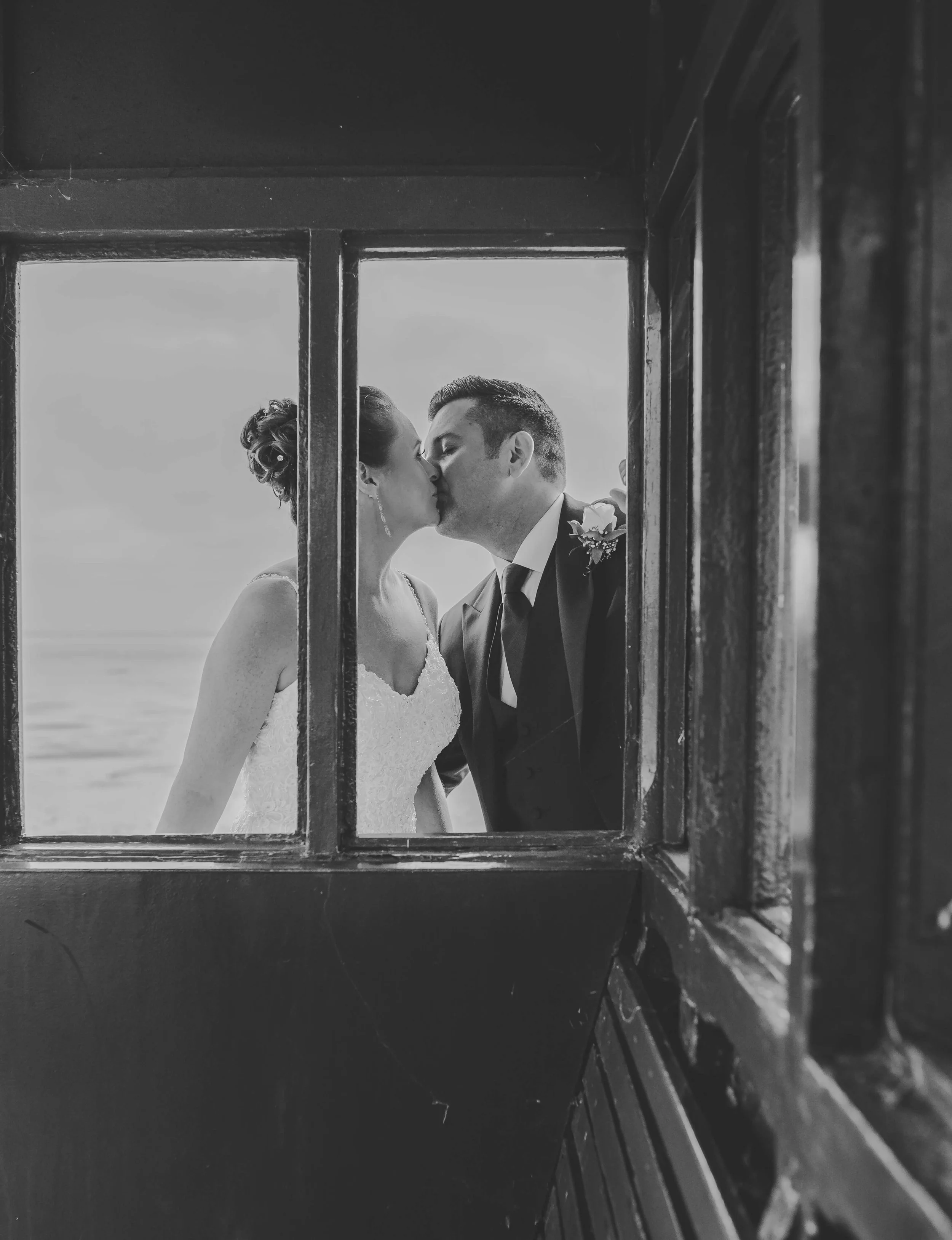 A black and white photo of a couple kissing, seen through a window frame, with the background suggesting a beach or open landscape.