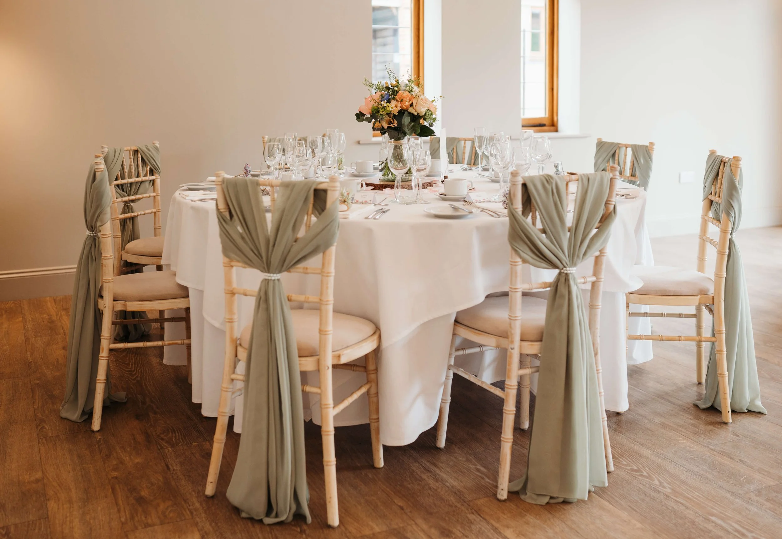 Round banquet table with a white tablecloth, set with multiple wine glasses, plates, and cutlery. A large floral centerpiece with peach and pink flowers in a glass vase decorates the middle. Eight chairs with beige cushions and green fabric tied arou