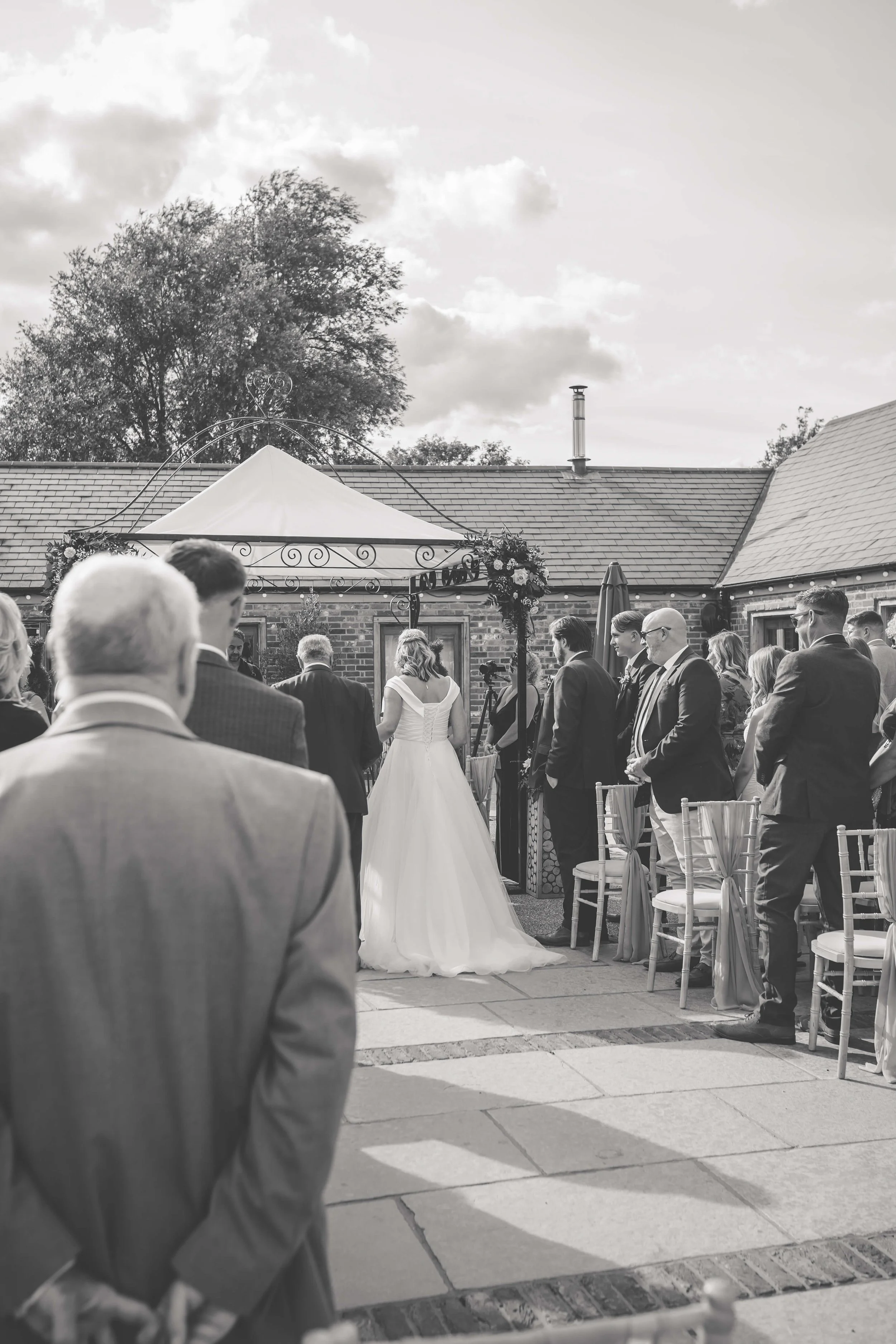 A black and white photo of an outdoor wedding ceremony with guests facing a bride and groom under a decorated arch.
