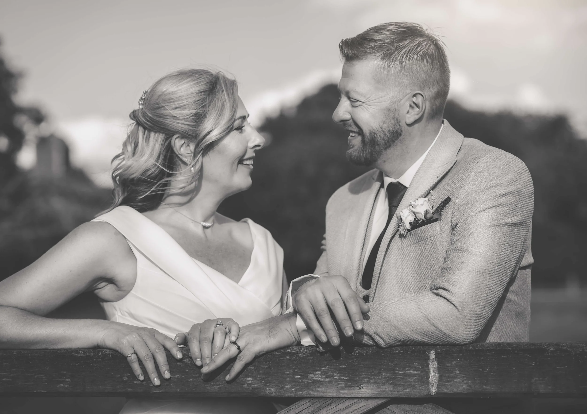 Black-and-white photo of a couple smiling and holding hands, standing outdoors with blurred trees and sky in the background.
