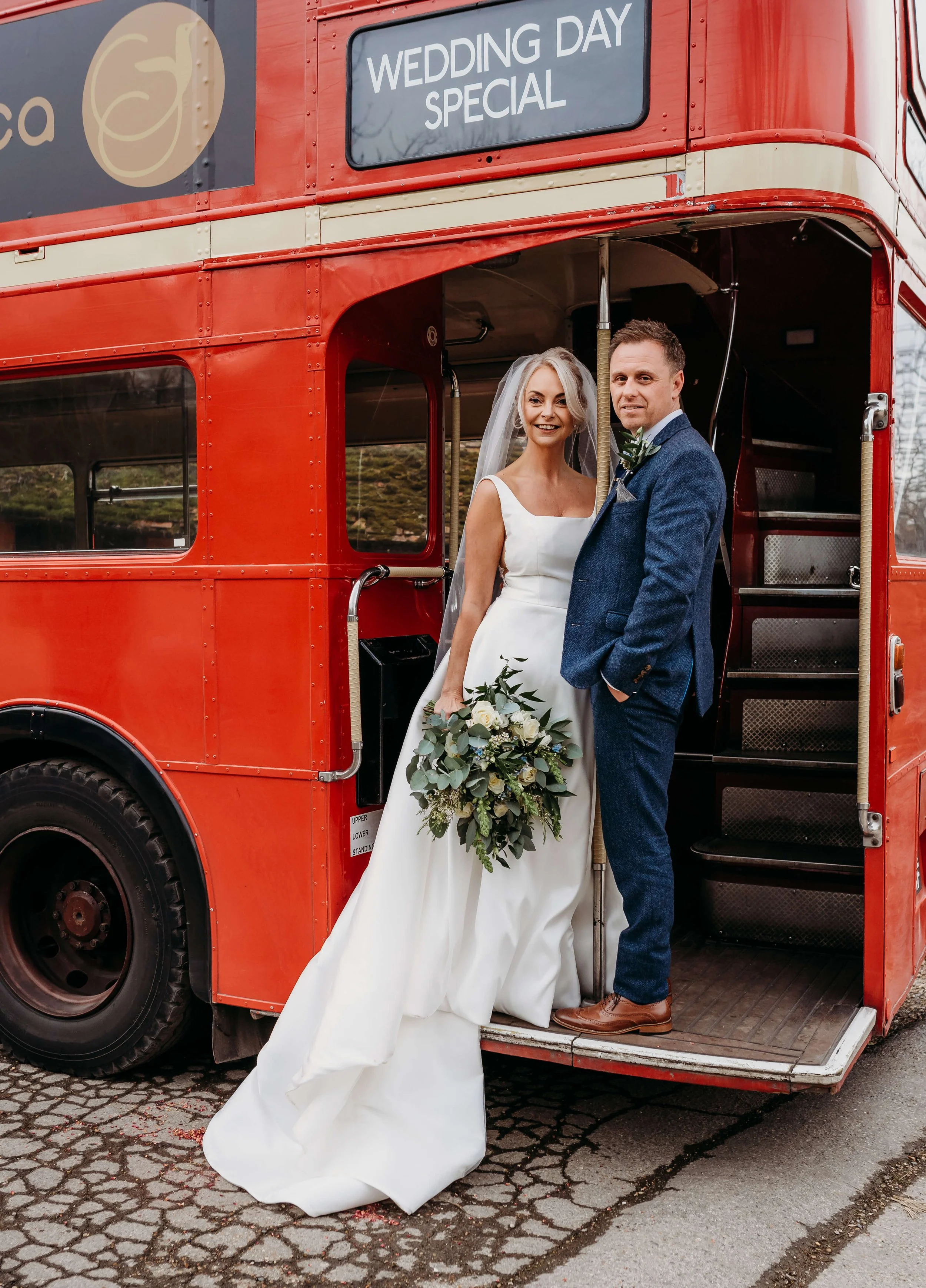 A bride and groom standing inside the open steps of a red double-decker bus, with a sign above that reads "Wedding Day Special". The bride is wearing a white wedding gown and holding a bouquet, and the groom is wearing a blue suit with a boutonniere.