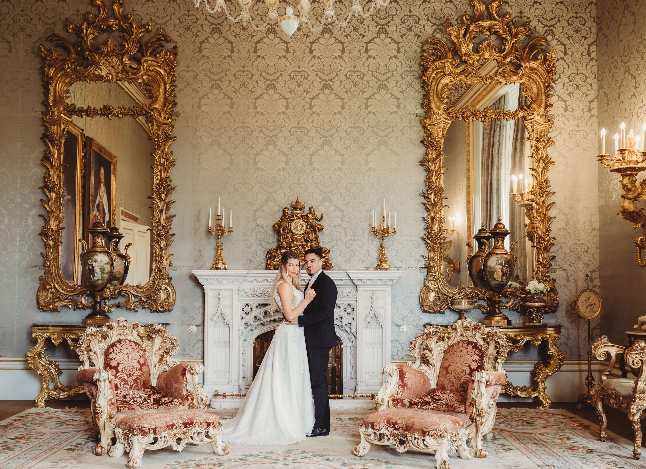 A bride and groom standing in an ornate, luxurious room with gold accents, antique furniture, and decorative mirrors.