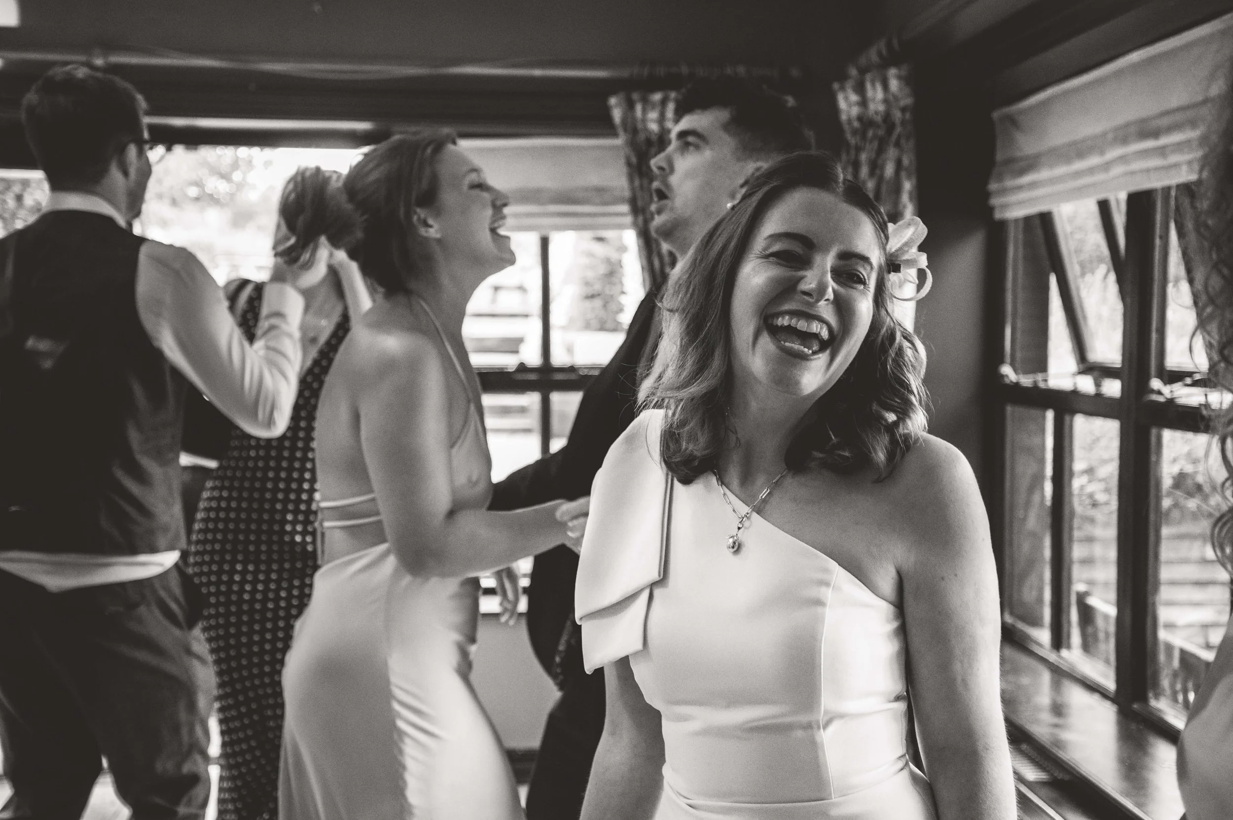 A group of people at a social gathering, with women in elegant dresses and men in suits, inside a room with large windows, smiling and laughing.