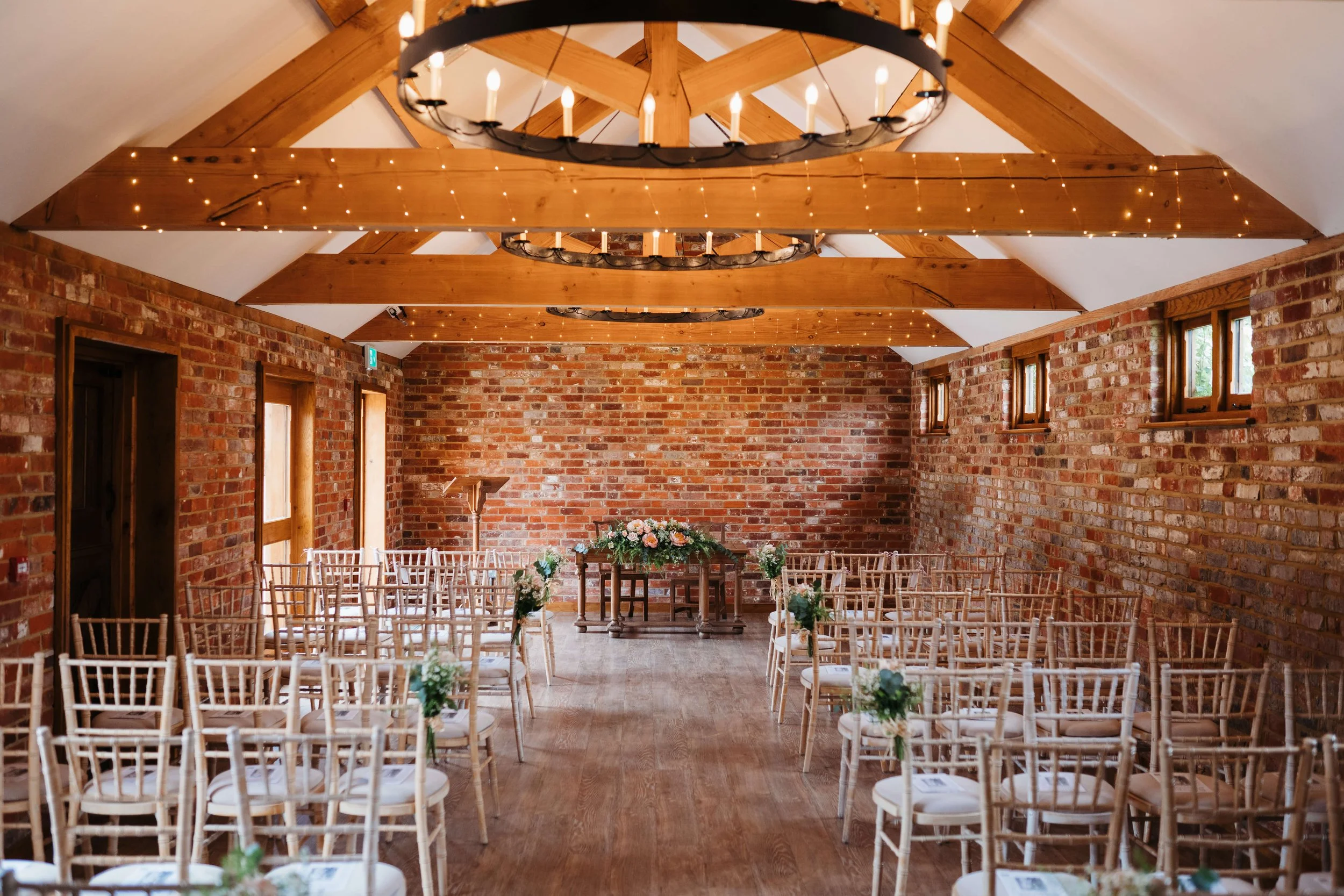 Wedding ceremony setup in a rustic indoor venue featuring brick walls, wooden beams, chandeliers, string lights, and rows of chairs decorated with flowers.