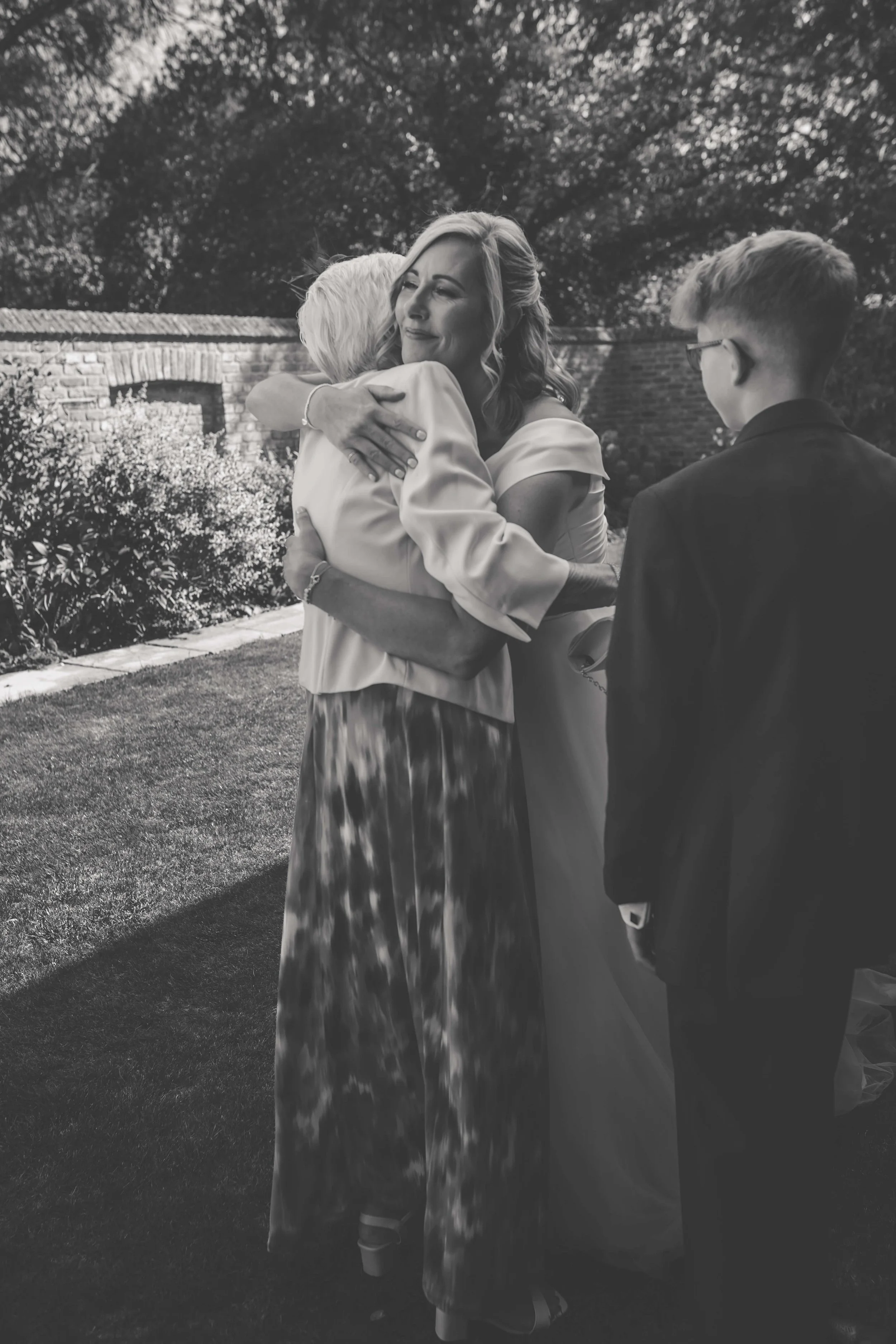 A woman in an off-shoulder dress embraces an elderly woman outdoors during a gathering, with a young man in a suit observing.