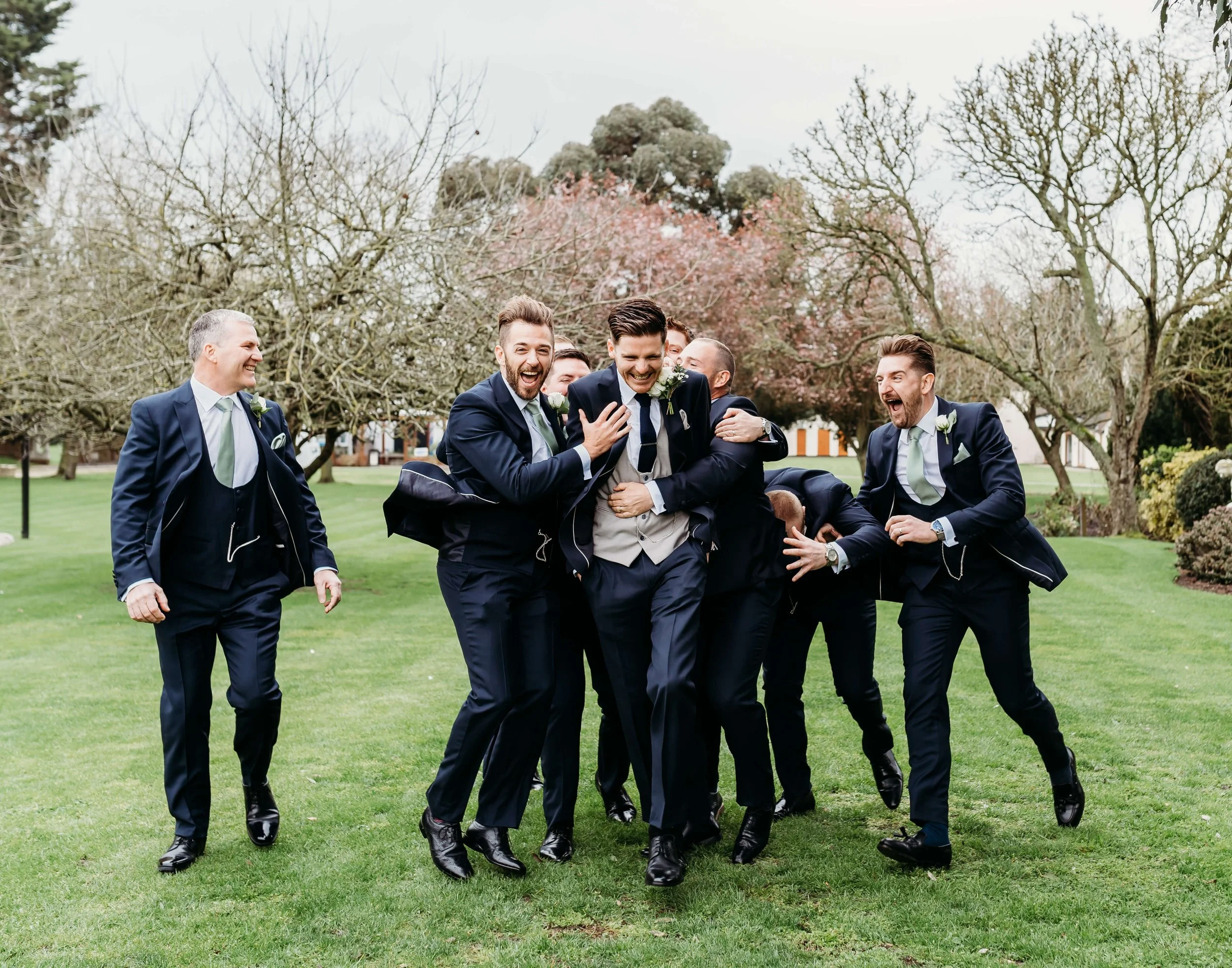A group of seven men in suits joyfully celebrating outdoors, with trees and grass in the background.