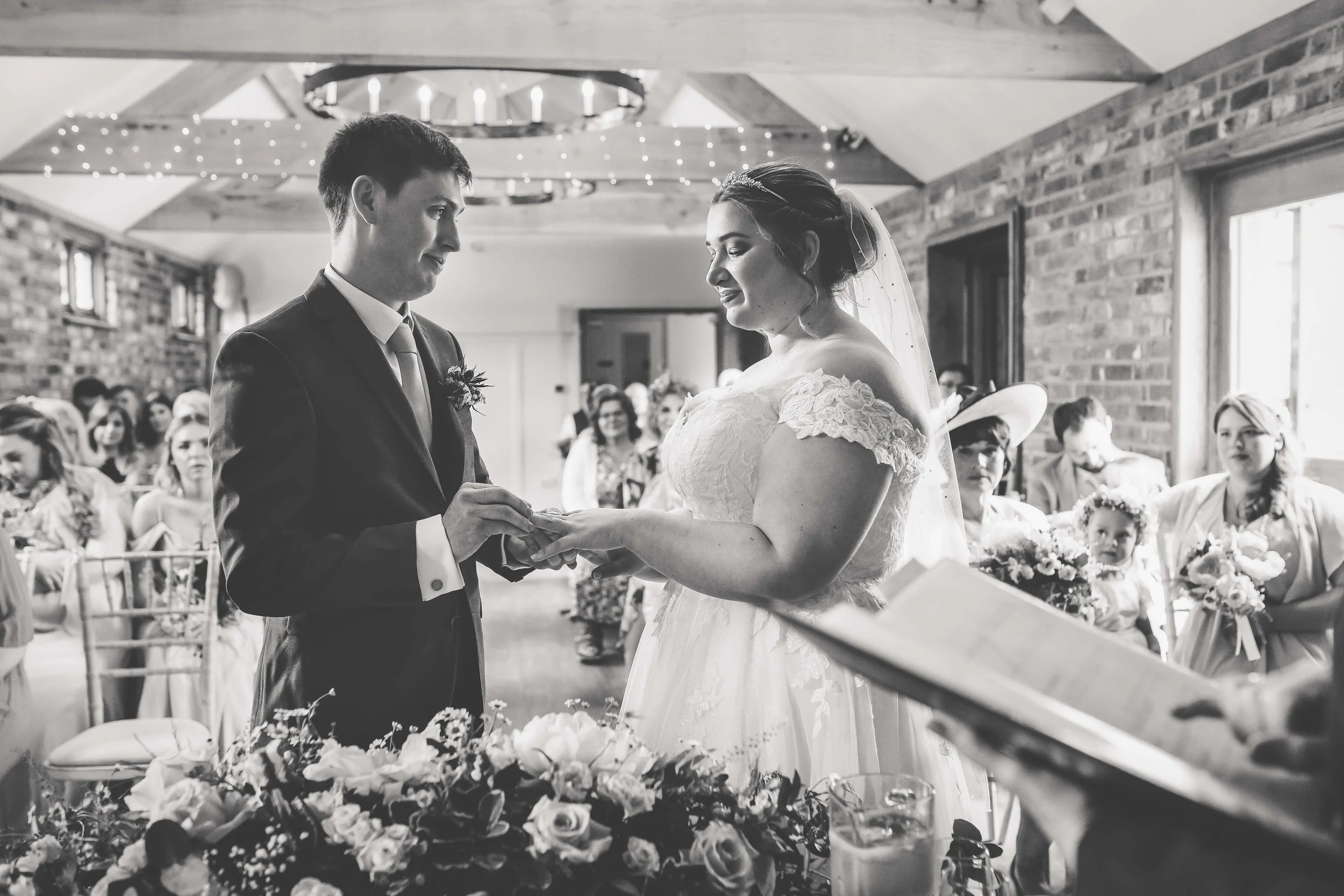 A couple getting married, exchanging rings in a wedding ceremony with guests seated in the background, inside a rustic venue with exposed brick walls and wooden beams.