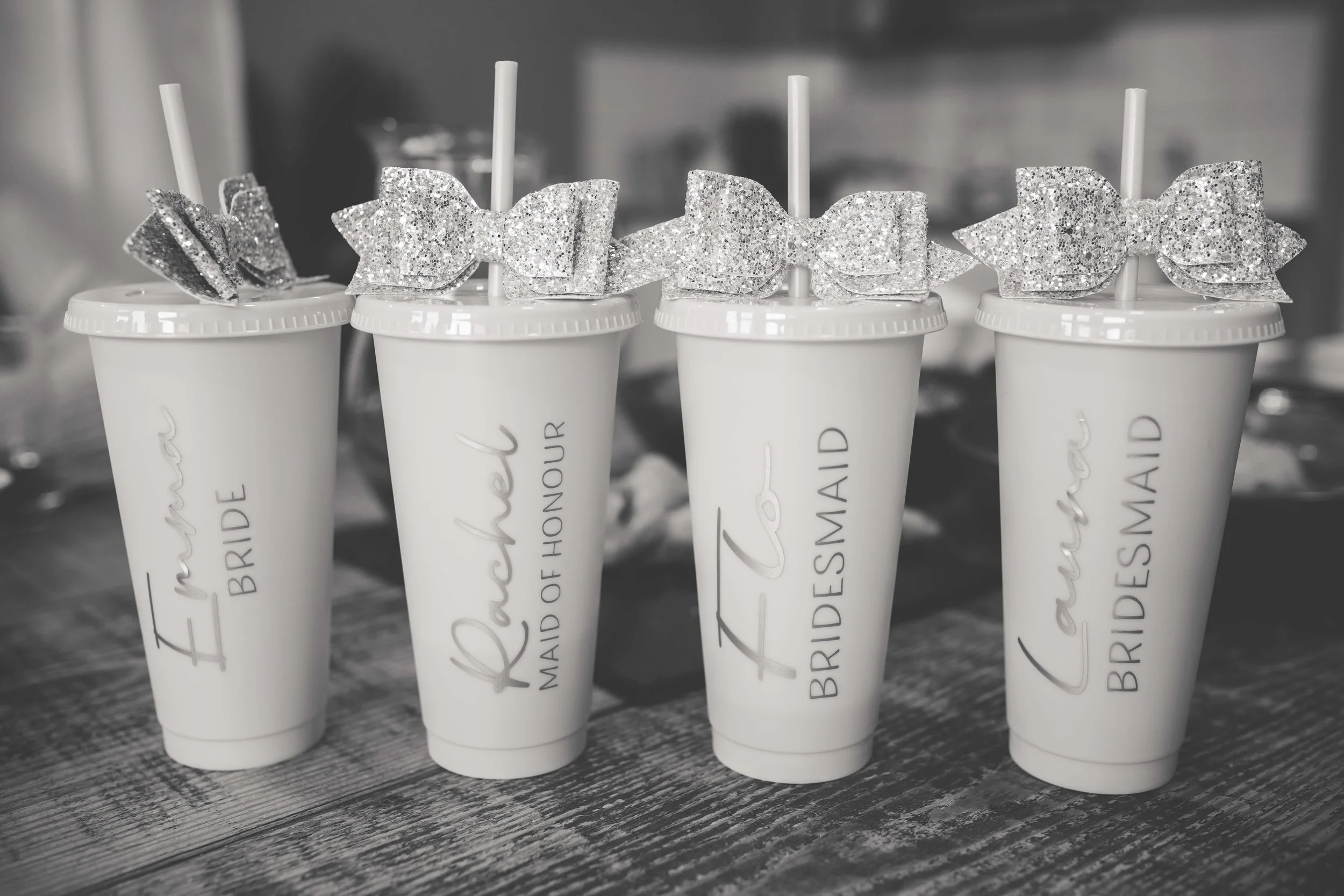 Four disposable cups with straws and glittery bows on top, each labeled with different wedding titles: bride, maid of honor, bridesmaid, and bride's maid, arranged on a rustic wooden table.