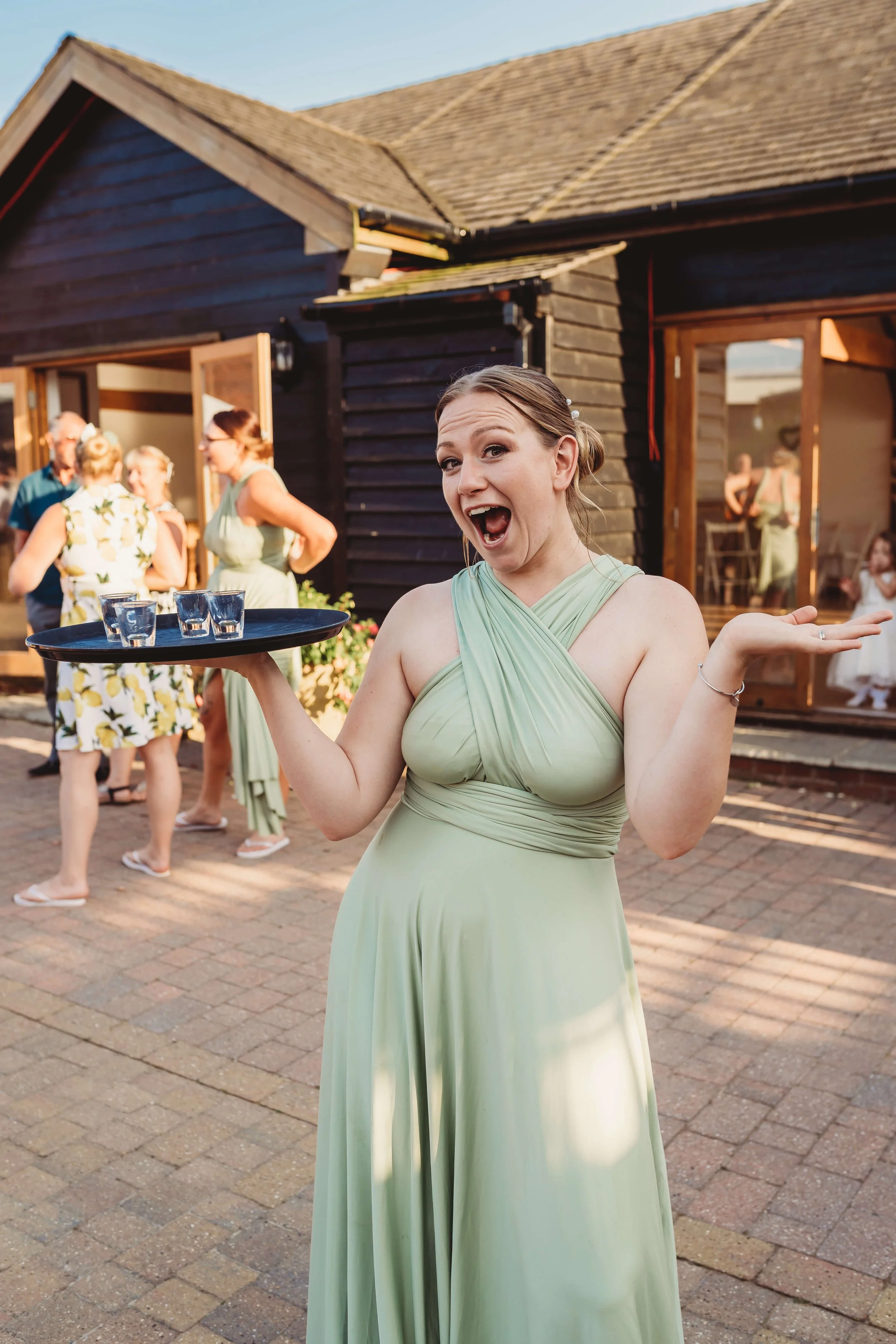 A woman in a light green dress holding a tray with three shot glasses, smiling and gesturing at a gathering outside a house.