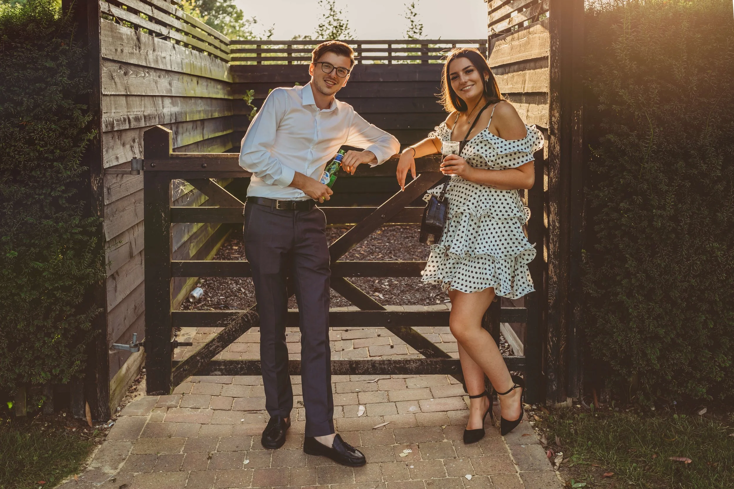 A man and woman standing outside near a wooden gate, both dressed formally, holding drinks and smiling at the camera. The man is wearing a white shirt, dark pants, and glasses, while the woman is wearing a polka-dot dress and high heels.