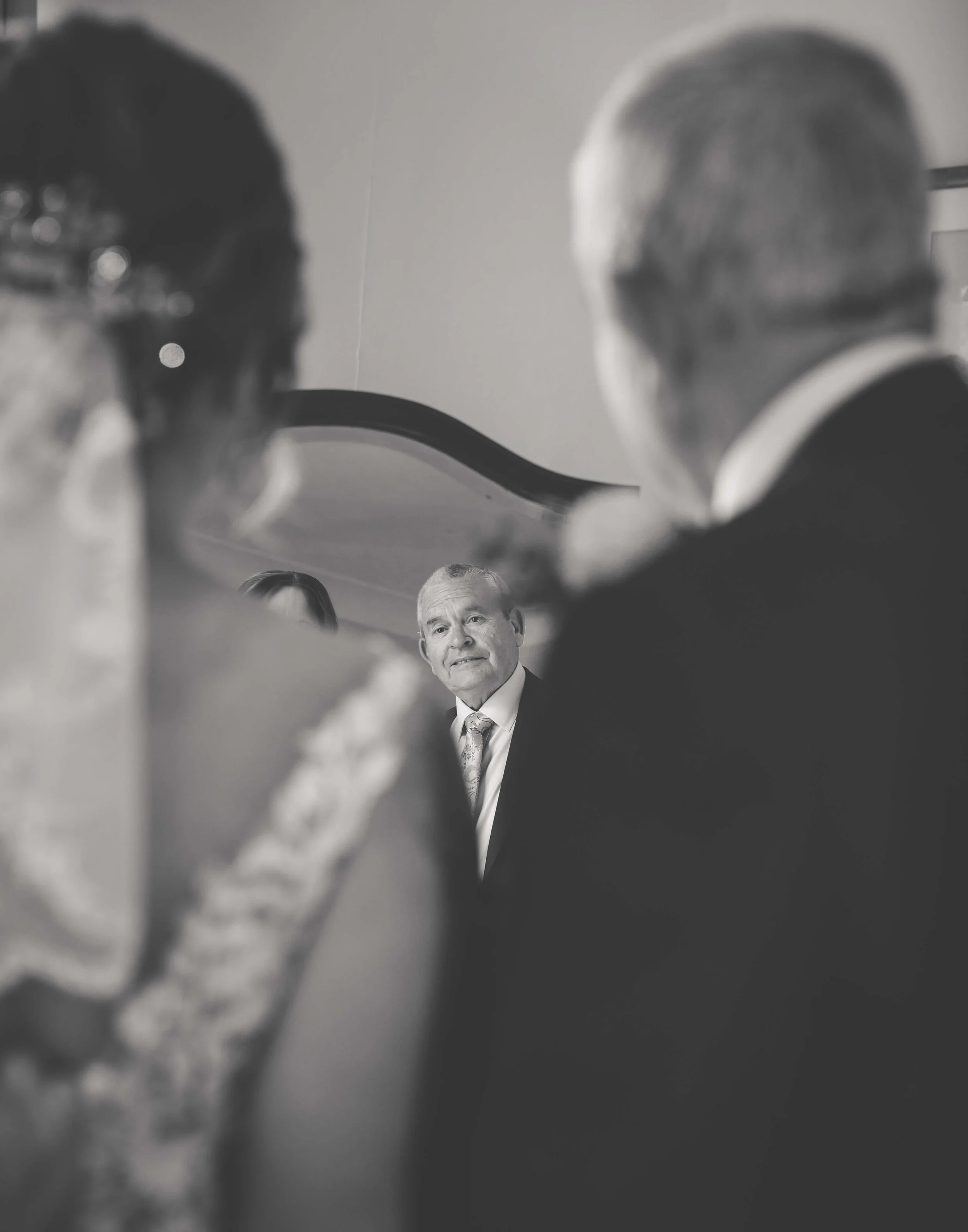 Black and white photo of a formal event with people in formal attire, focusing on a man in the background who is looking at the camera, while the others are blurred in the foreground.