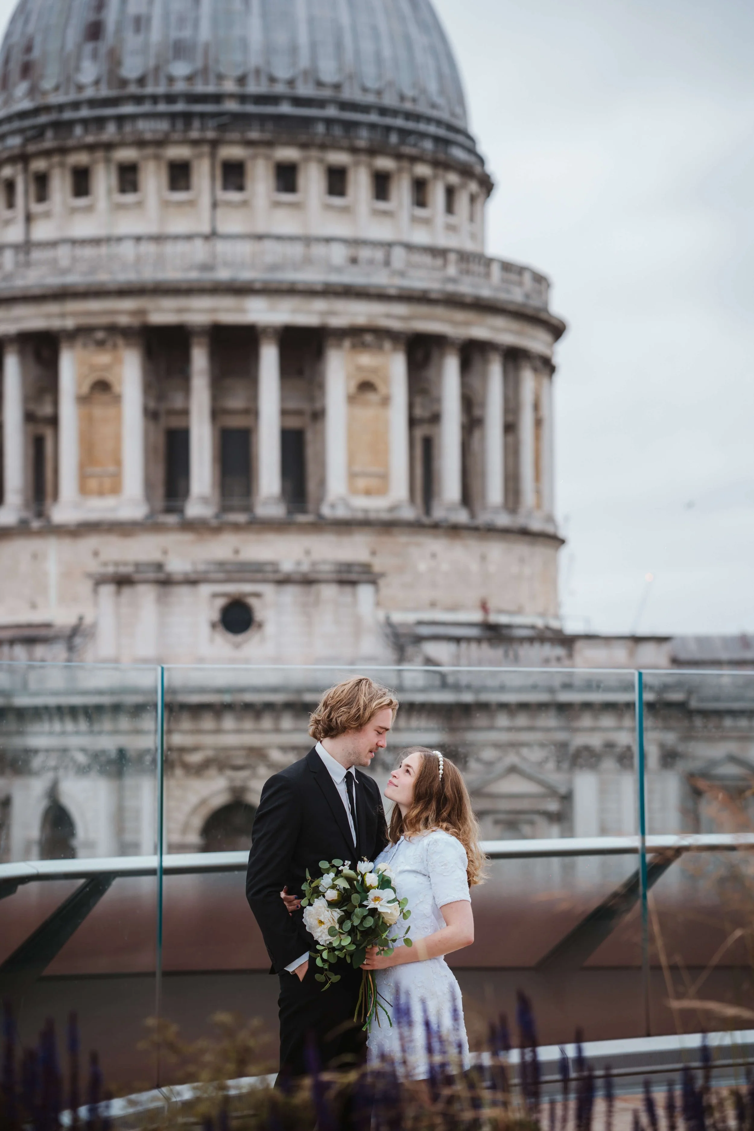 A couple in wedding attire, the groom in a black suit and the bride in a white dress, holding a bouquet of flowers, standing on a rooftop with the dome of St. Peter's Basilica in the background.
