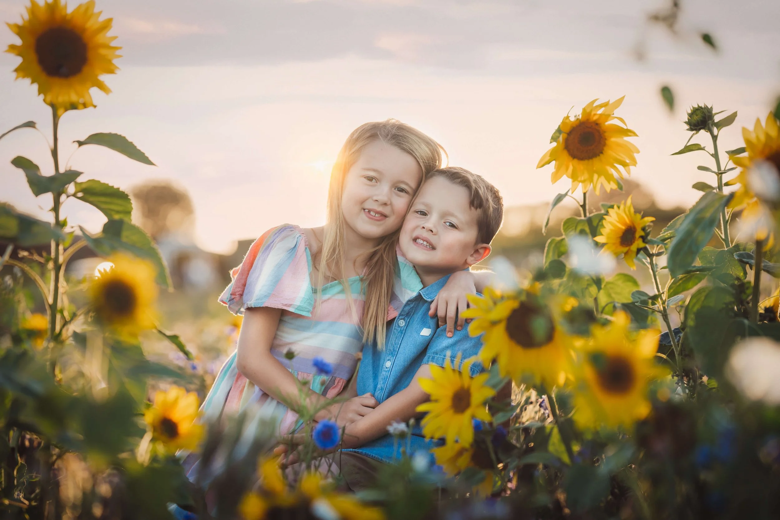 Two children, a girl and a boy, smiling and hugging each other in a sunflower field during sunset.