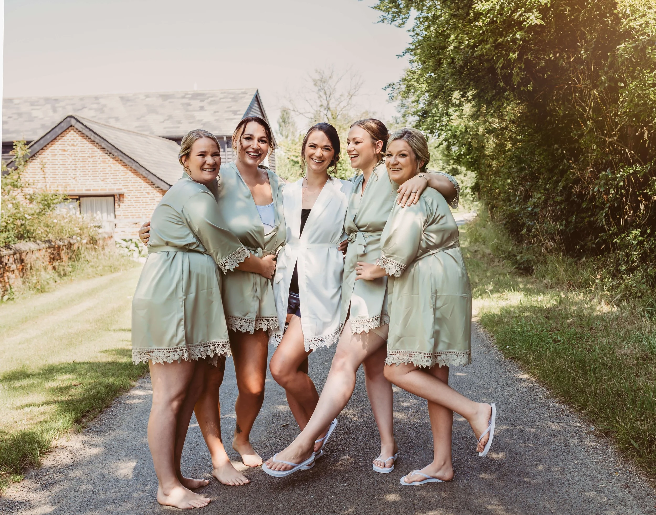 Six women, five in matching satin robes and one in a white robe, are standing close together outside on a gravel path, smiling and having fun on a sunny day with a house and trees in the background.