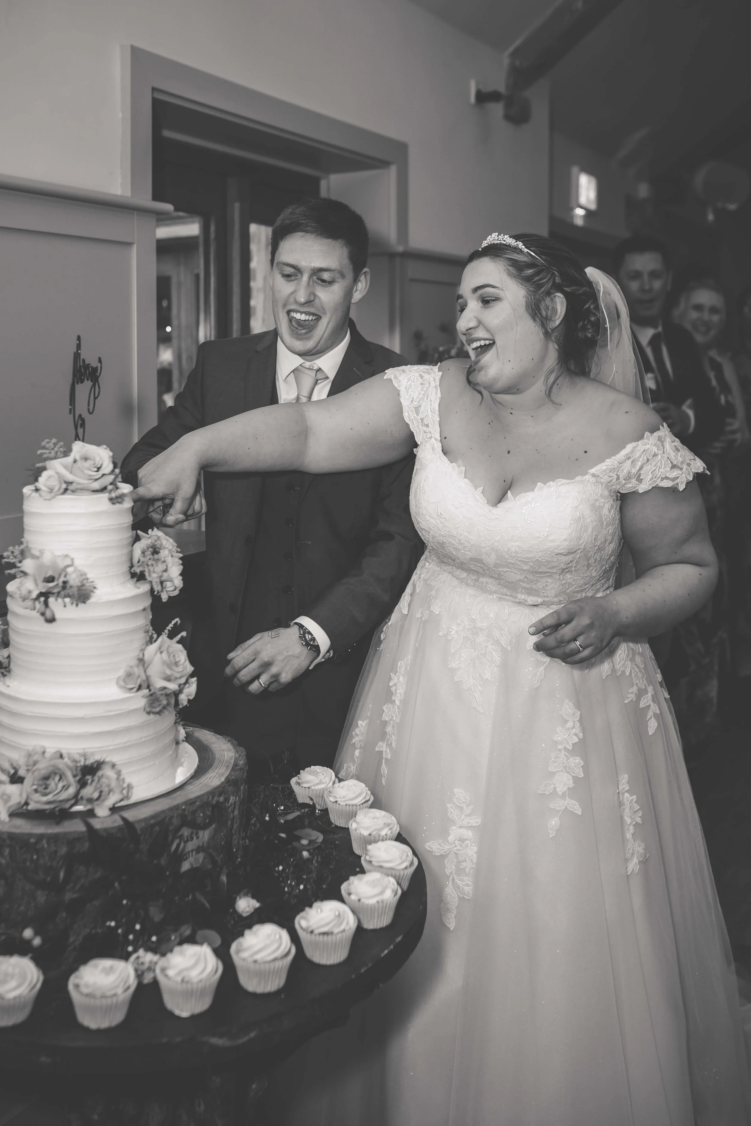 A black and white photo of a bride and groom cutting their wedding cake together, surrounded by guests.