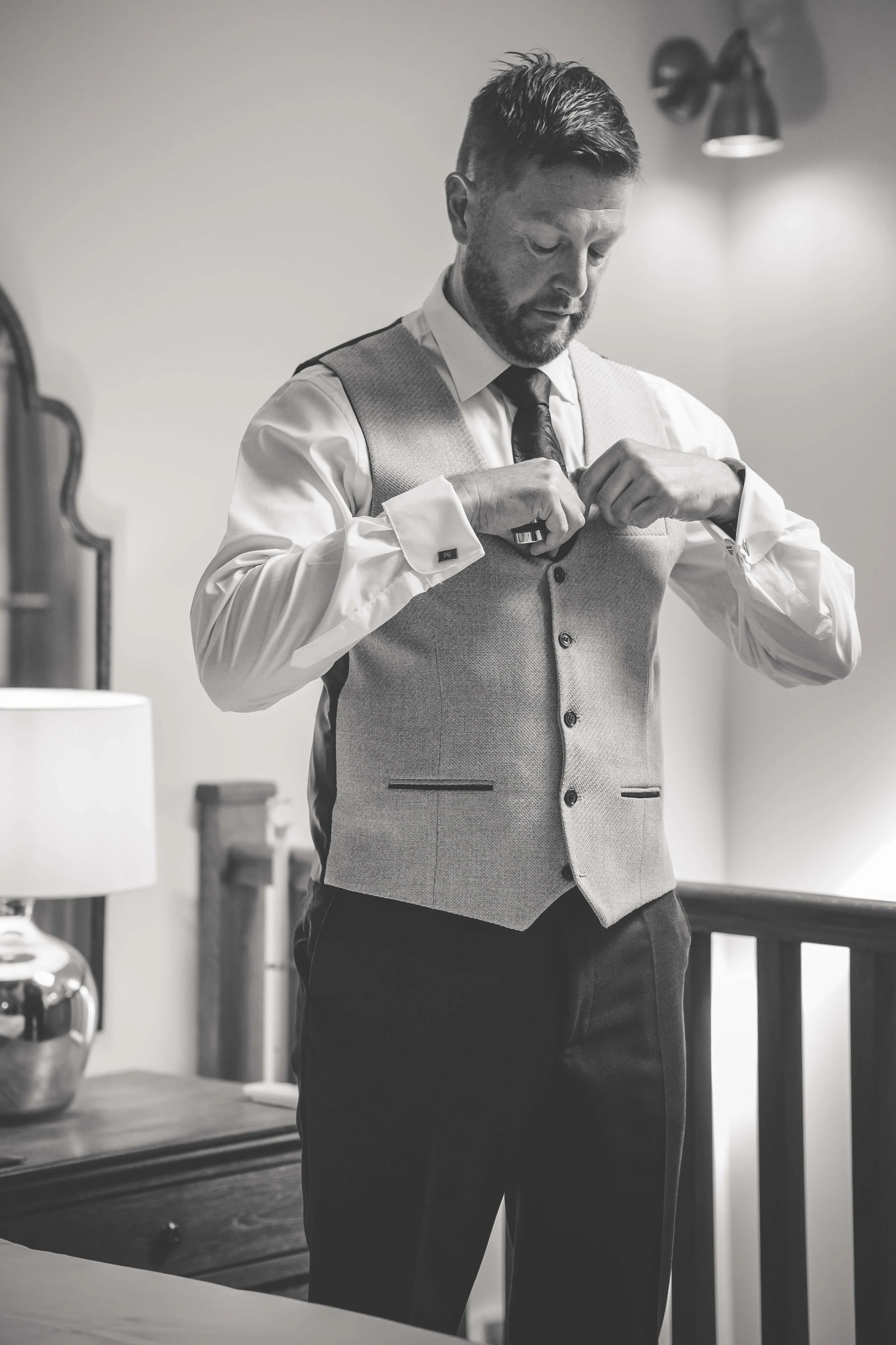 A man in formal attire, adjusting his suit jacket in a room with a lamp and mirror.