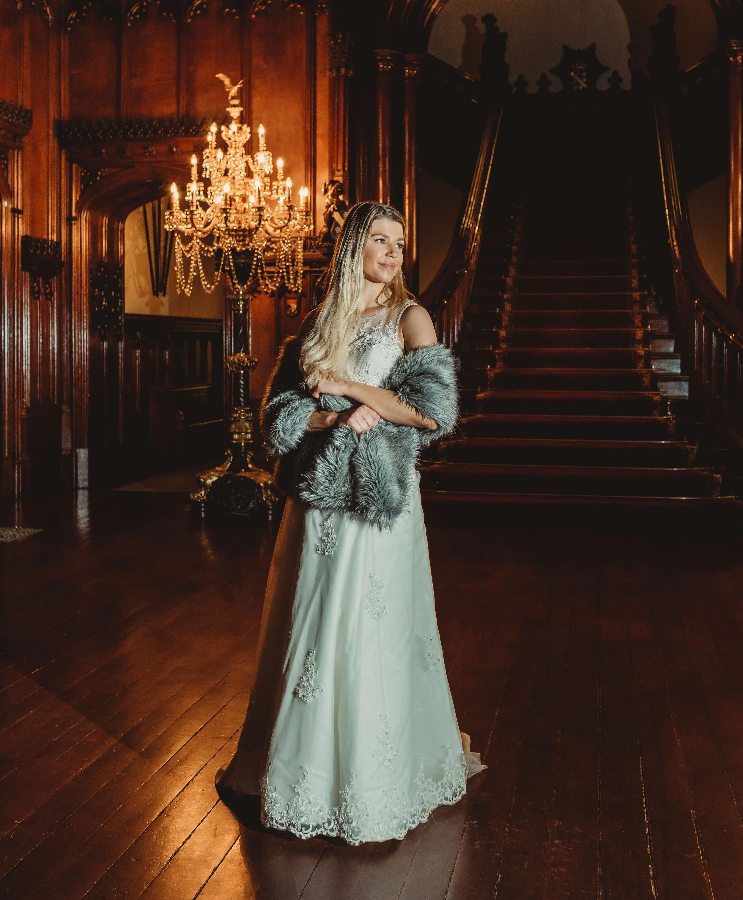 A young woman in a white wedding dress with lace details, wearing a gray fur stole, stands in a grand, wood-paneled hall with a large staircase and a chandelier.