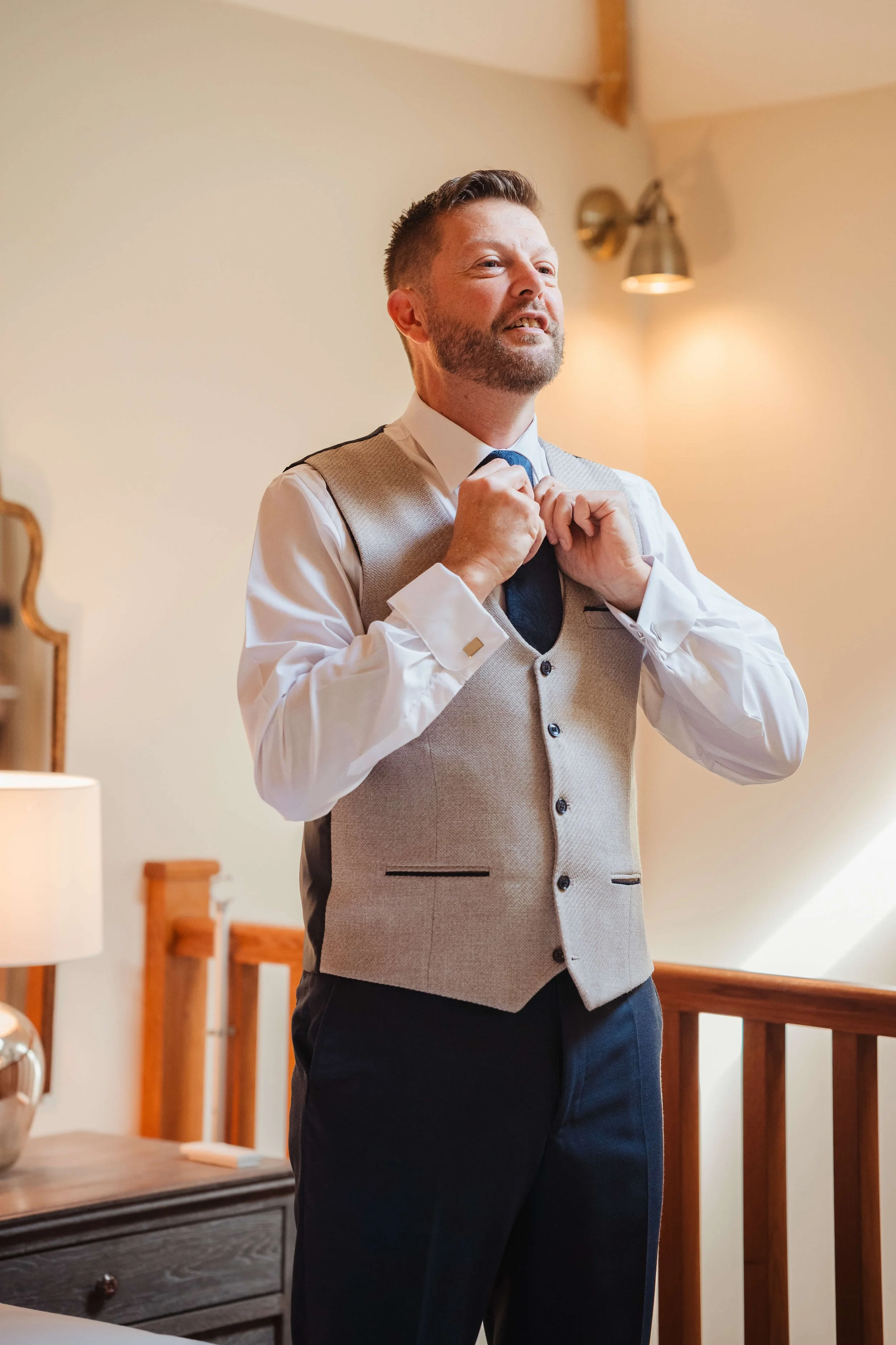 A man adjusting his tie in a warmly lit room, dressed in formal attire including a vest, white shirt, and dark pants.