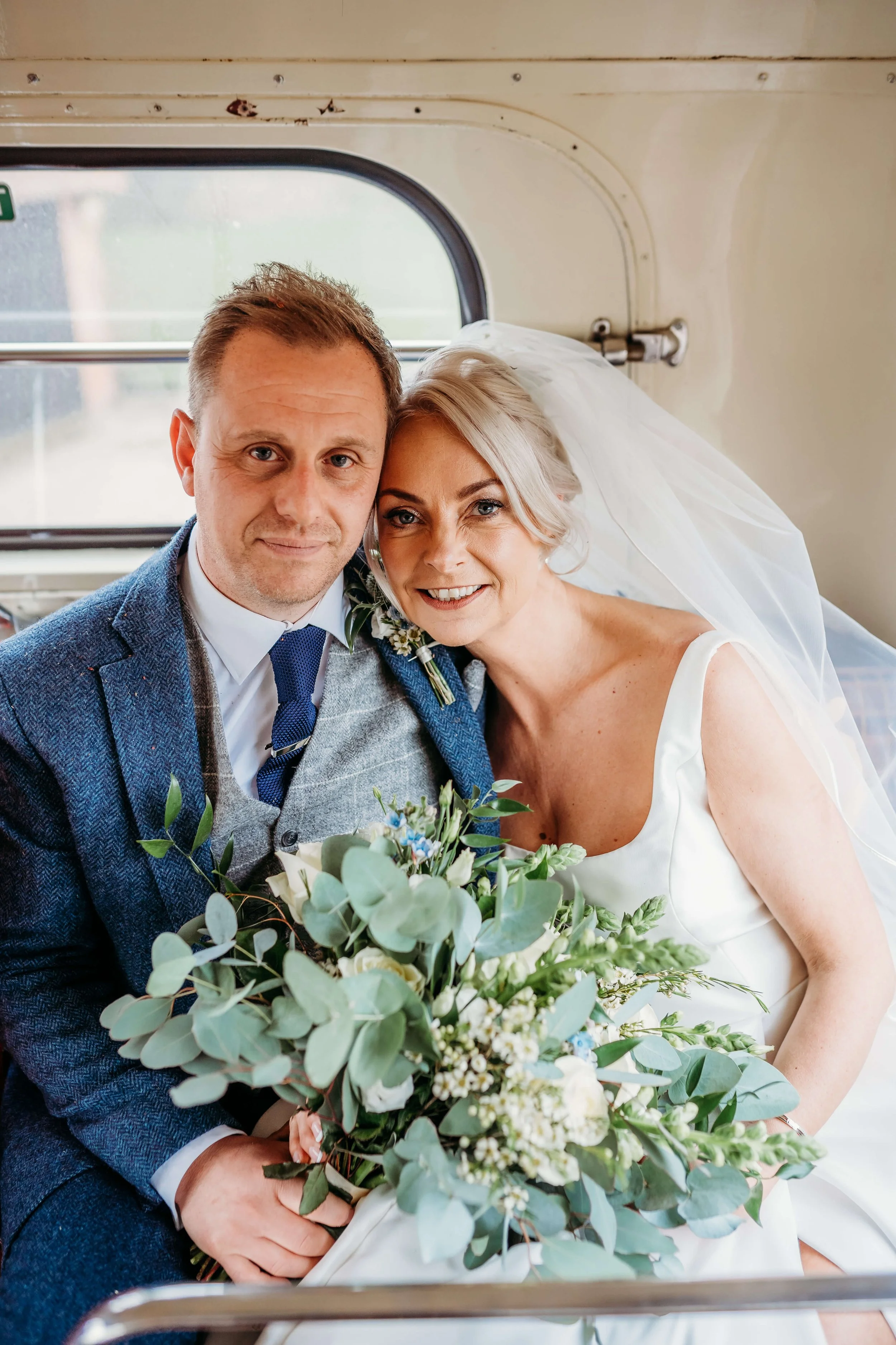 A newlywed couple sitting close together inside a train, smiling at the camera. The bride has a bouquet of flowers, and the groom is dressed in a blue suit with a tie.