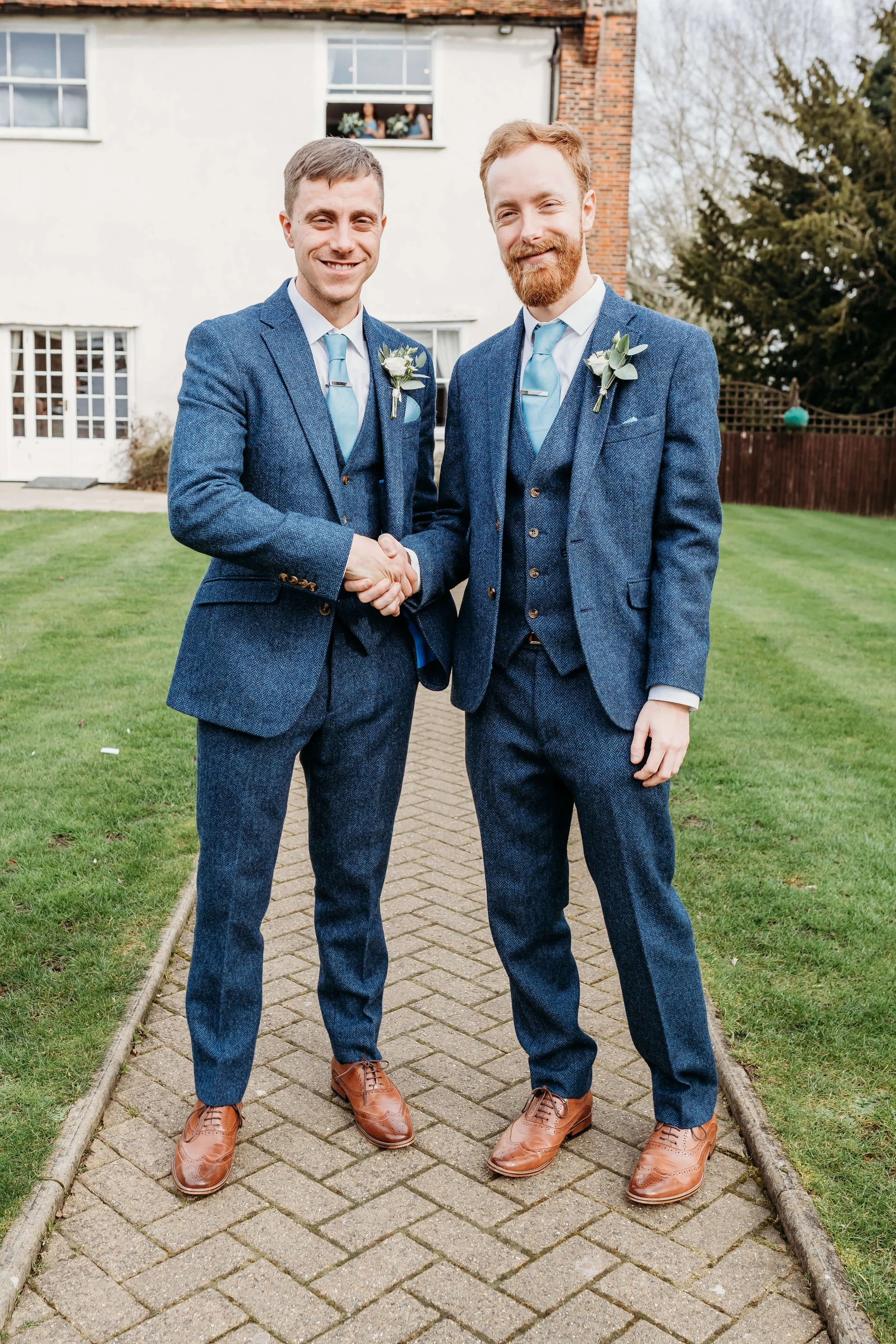 Two men in blue suits shaking hands outdoors, smiling, with a white house and trees in the background.